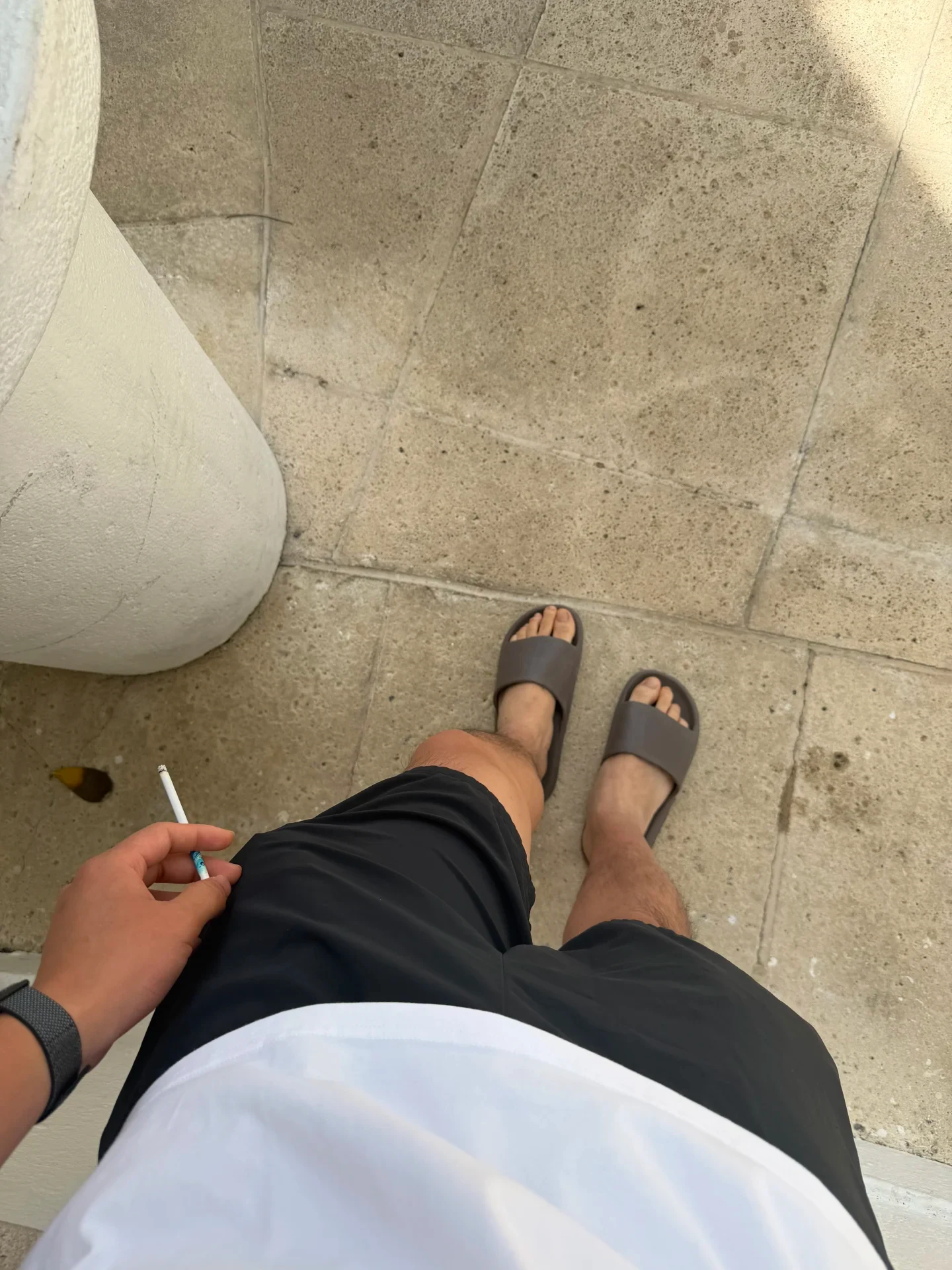 Top-down view of a person wearing black shorts, a white shirt, and slide sandals standing on a stone tile floor while holding a lit cigarette.