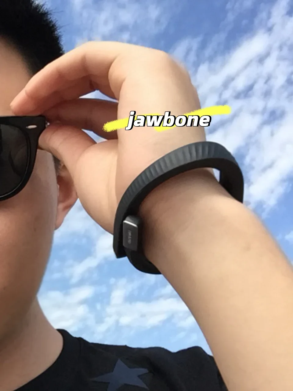 A person adjusting their sunglasses against a blue cloudy sky, wearing a black Jawbone fitness tracker band on their wrist, with the word 'jawbone' overlaid in text.