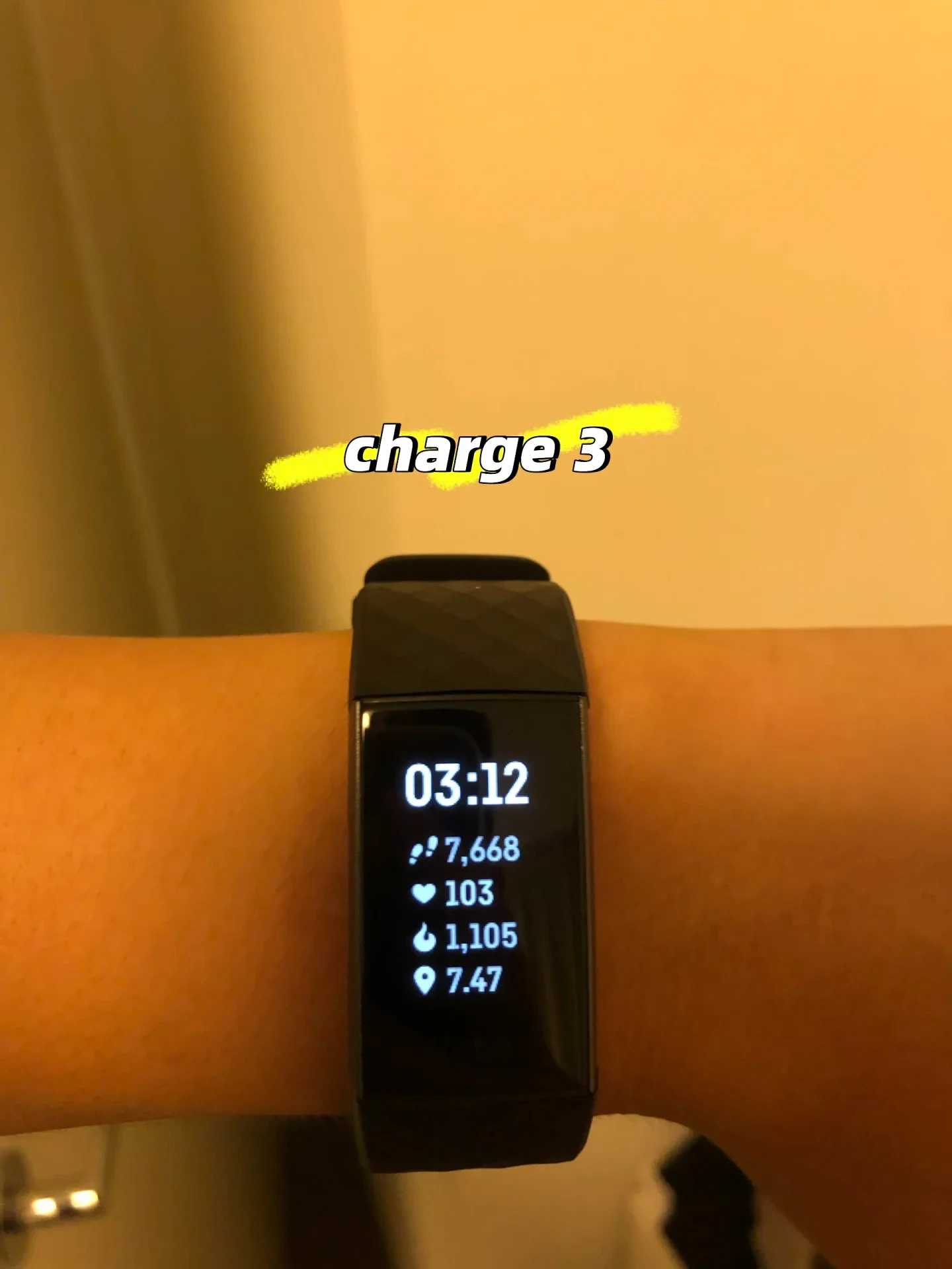 A Fitbit Charge 3 fitness tracker worn on a wrist displaying 03:12 alongside metrics for steps, heart rate, calories, and distance, with 'charge 3' written above it.