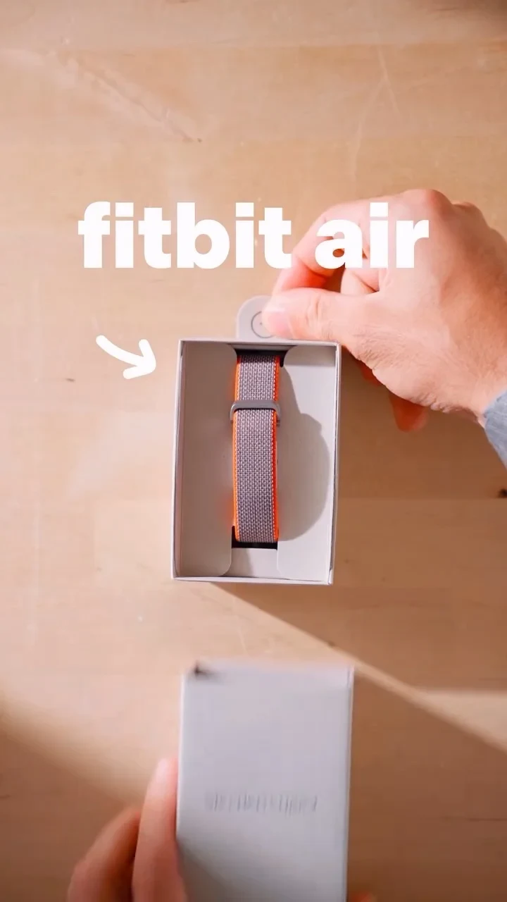 A person unboxing a grey woven watch band with orange edges, shown inside its opened packaging with overlay text reading 'fitbit air'.