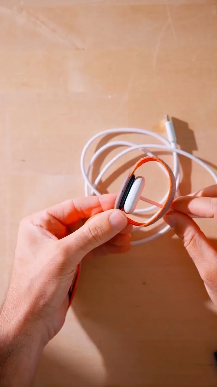 Two hands holding a smartwatch with an orange fabric loop strap attached to a magnetic charging puck, with a coiled white charging cable on a wooden surface in the background.