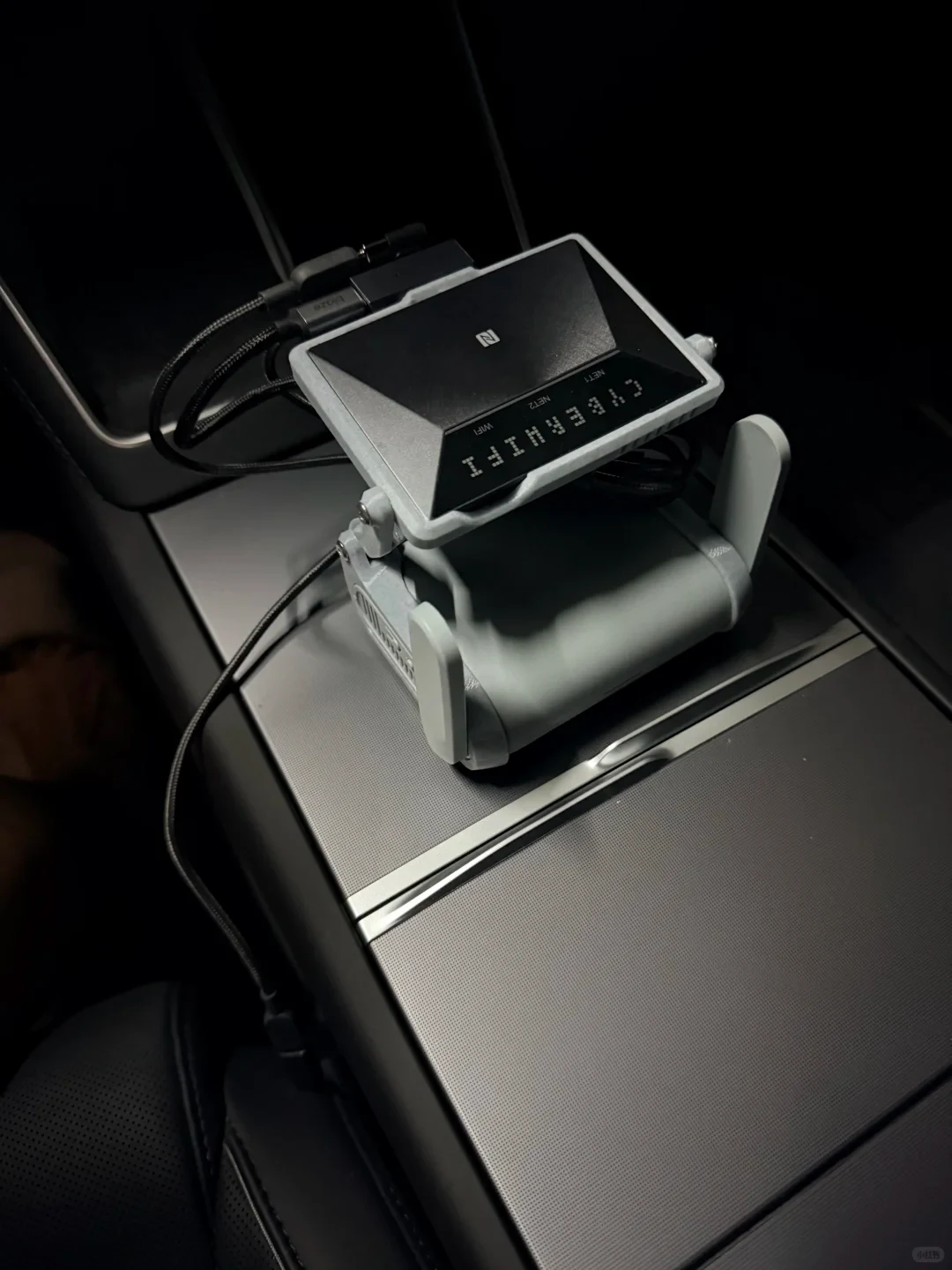 A gray electronic device with an angled display and attached cables rests on a car’s center console. Its screen shows a dot-matrix readout and small labels.