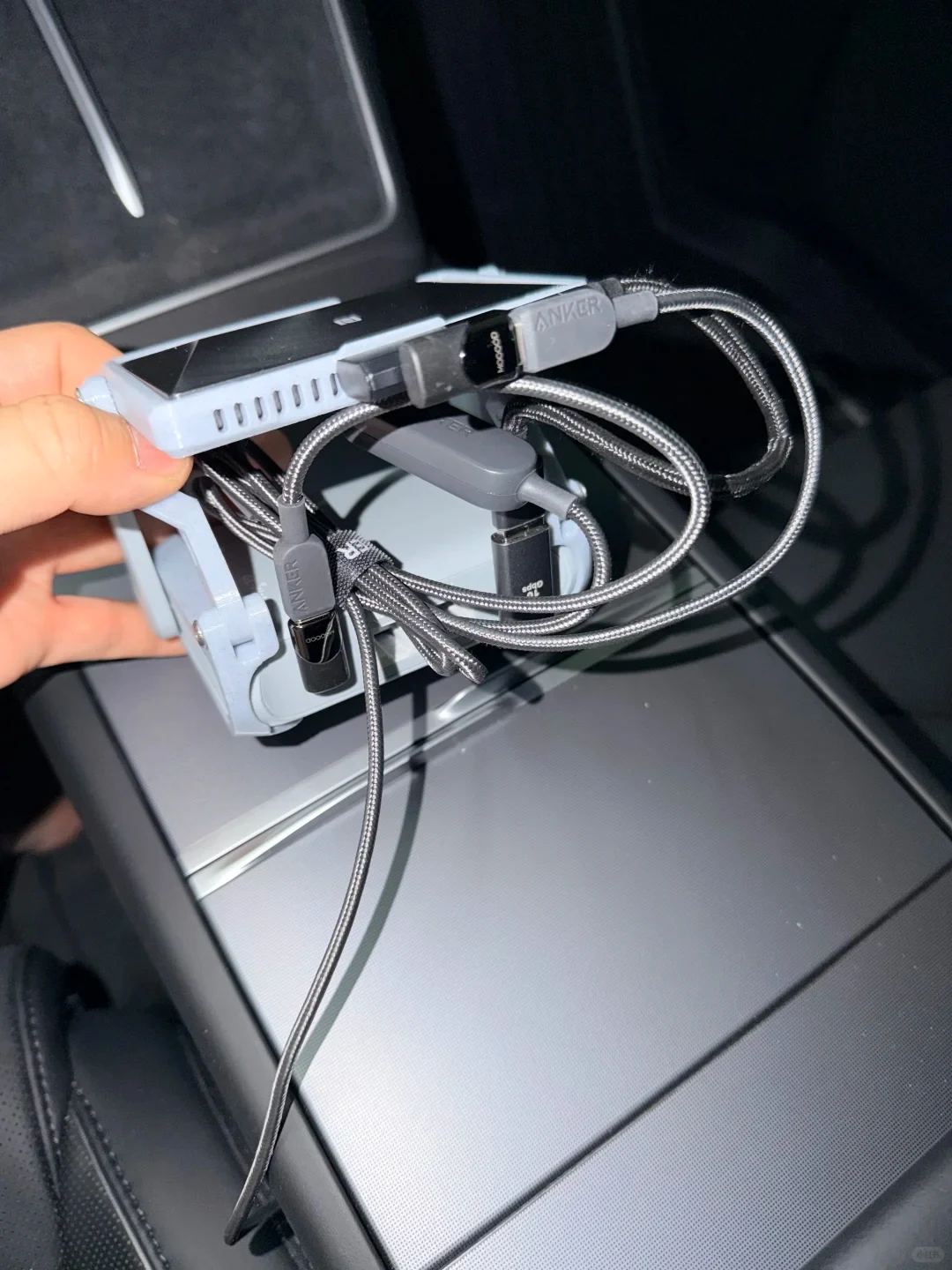 A hand holds a light-gray bracket with a black mobile Wi-Fi device stacked above a silver router, exposing braided Anker cables and adapters in the car’s center-console area.