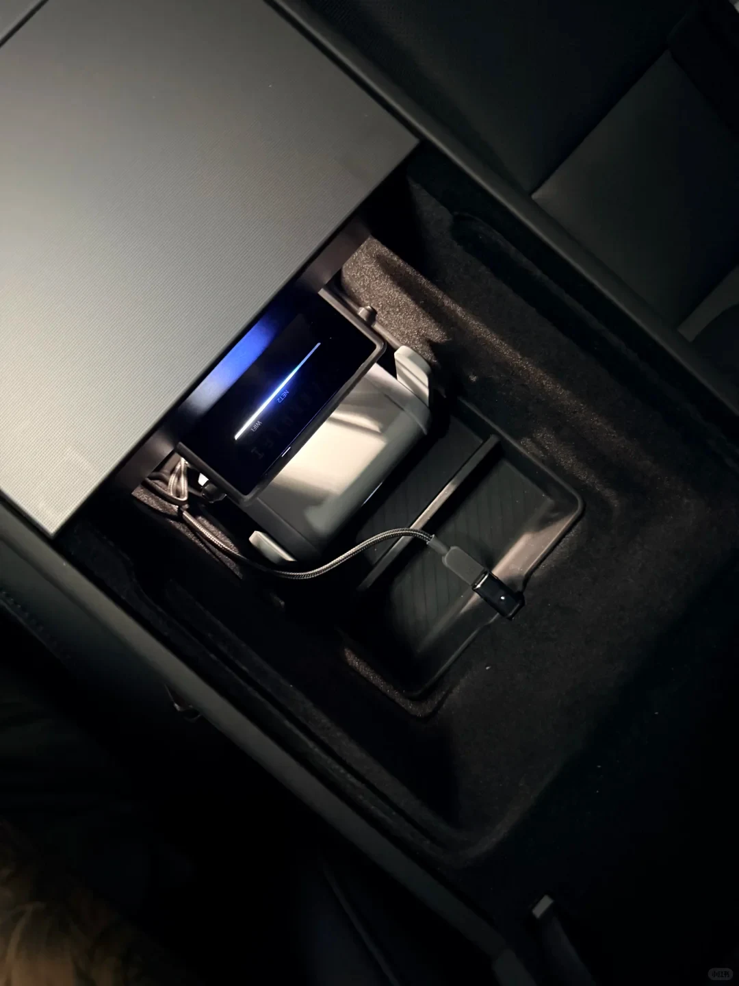 A photo of an open car-console compartment containing a small black electronic device with a glowing blue line on its display and a braided cable plugged in.