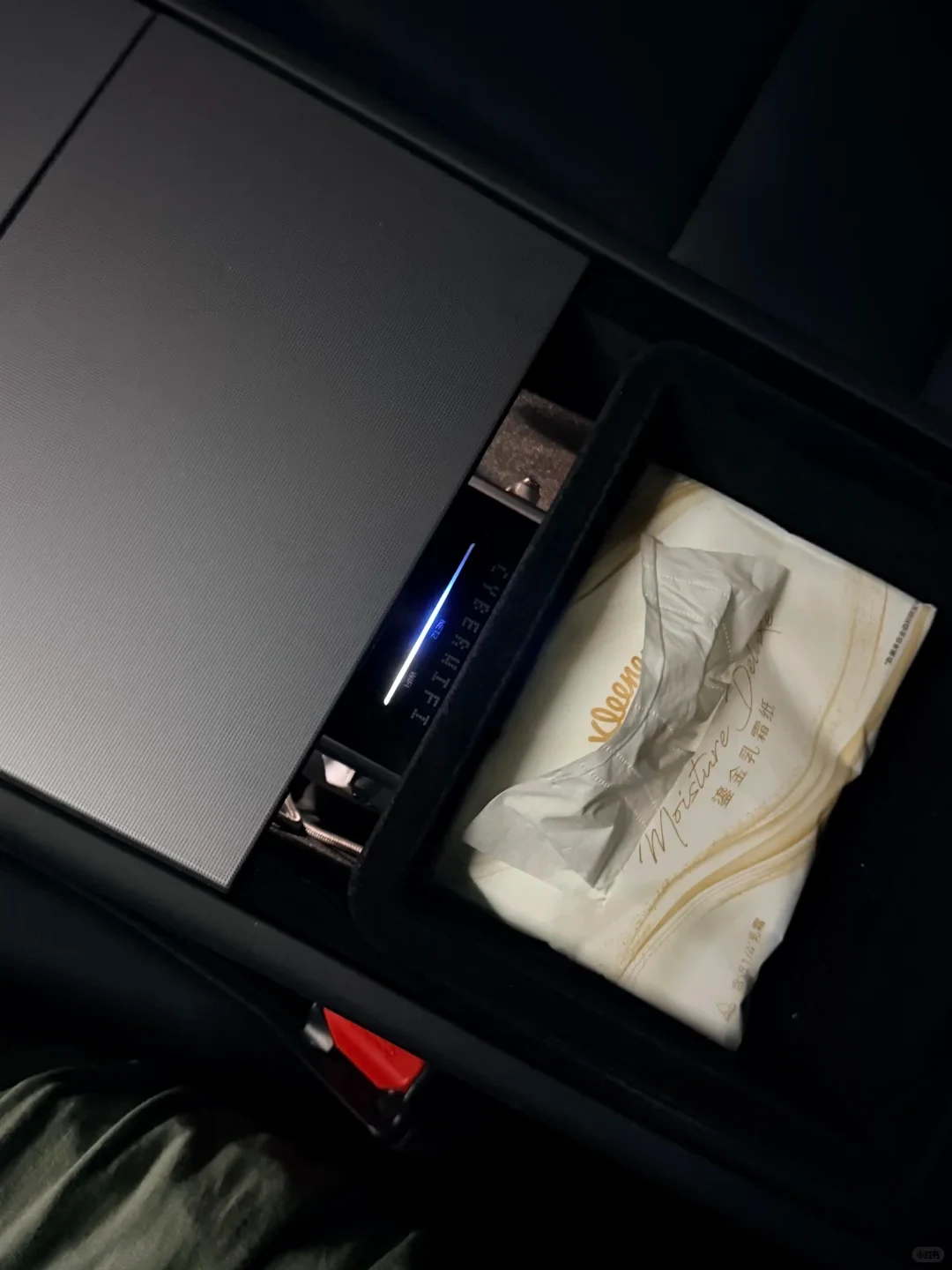 A powered black device is partly tucked beneath the car’s center-console lid beside a tissue packet, with its illuminated indicator bar visible.