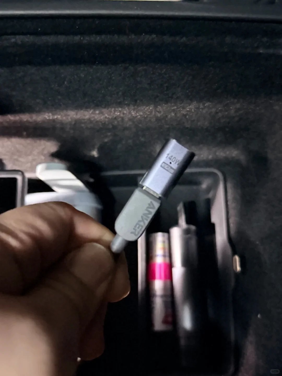 A hand holds a small gray USB adapter over an open storage compartment, with other small items inside.