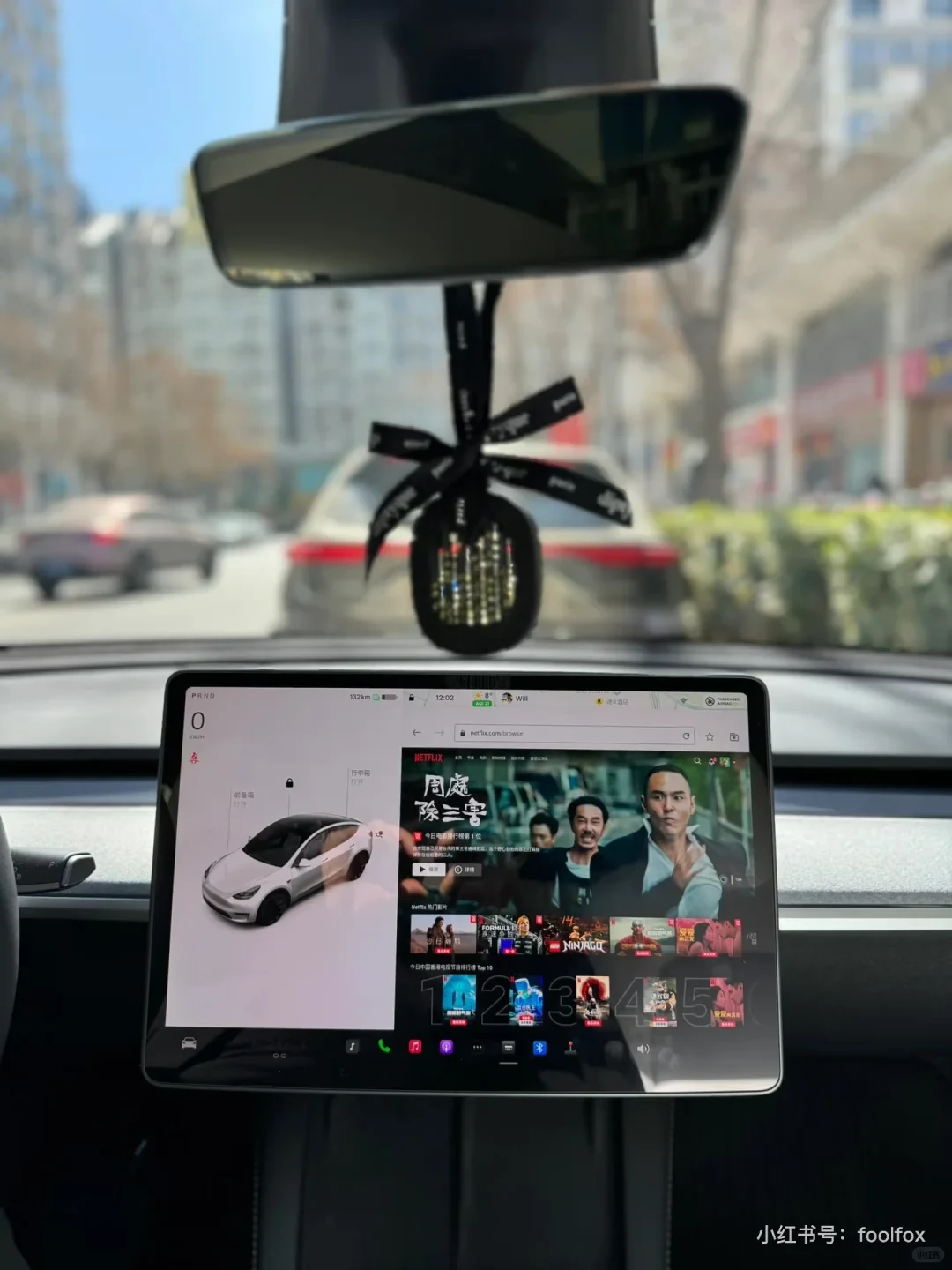 The car’s touchscreen shows a Netflix browsing page beside the vehicle controls while the speed display reads 0 km/h. A black ornament hangs from the rearview mirror, and cars and buildings are visible ahead.