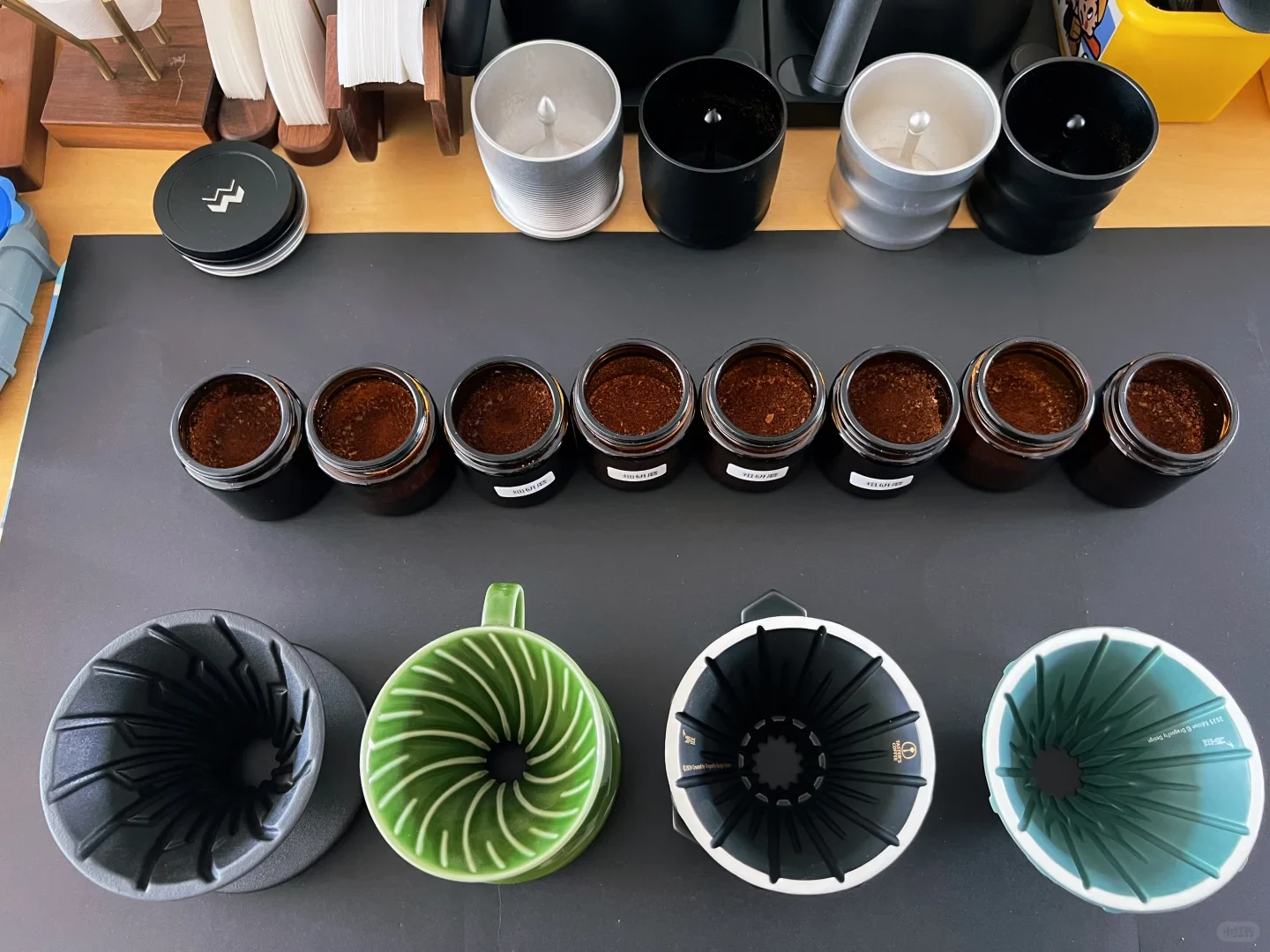A countertop display shows eight small open jars of ground coffee in a row, with several metal cups behind them and four large fluted coffee drippers in black, green, black-and-white, and turquoise in the foreground.