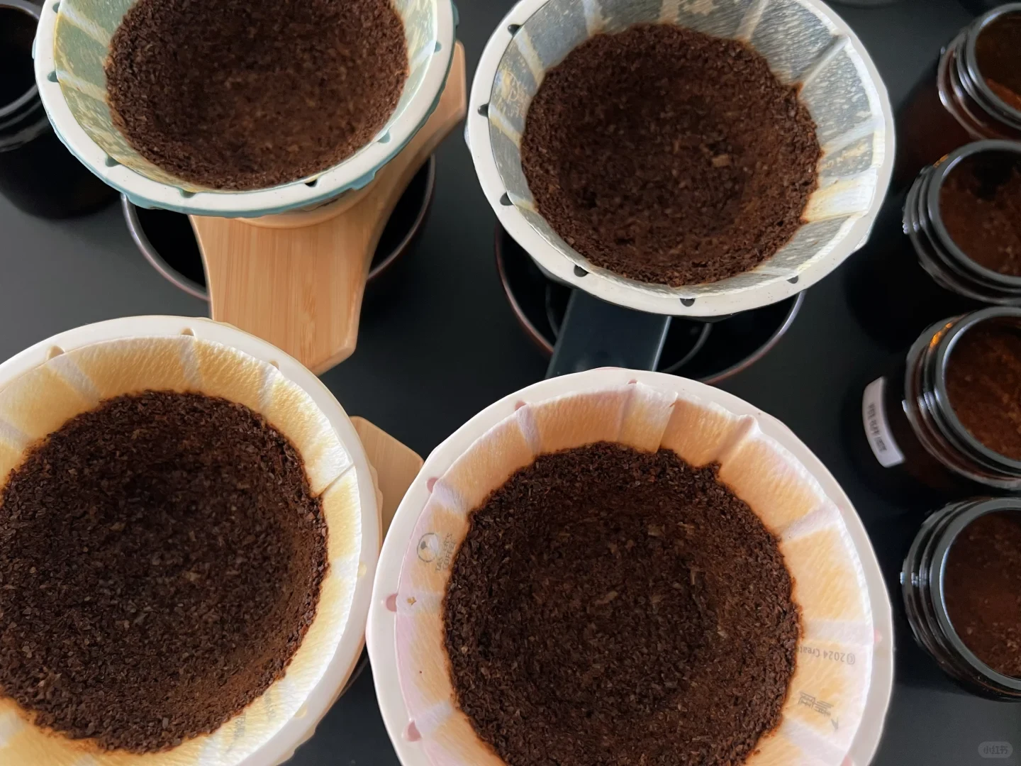 Overhead view of four paper coffee filters filled with dark, damp coffee grounds, arranged on a dark surface beside small jars.