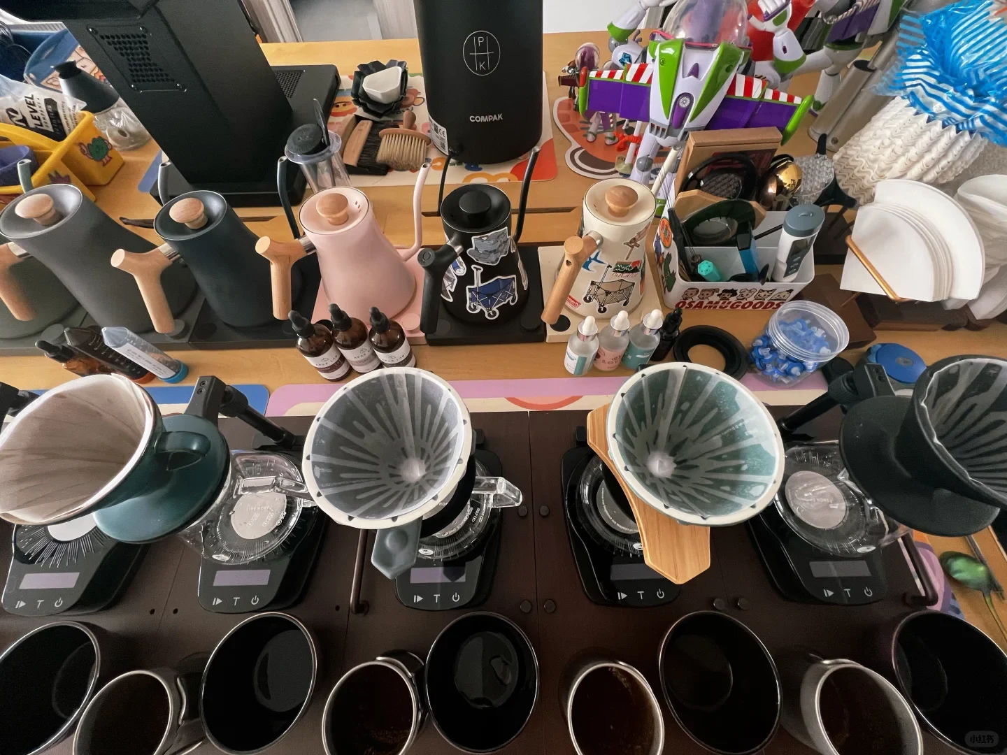 A crowded tabletop display of kitchen and household items, including several pour-over coffee drippers on digital scales, handled kettles, bottles, cups, and assorted toys and accessories.