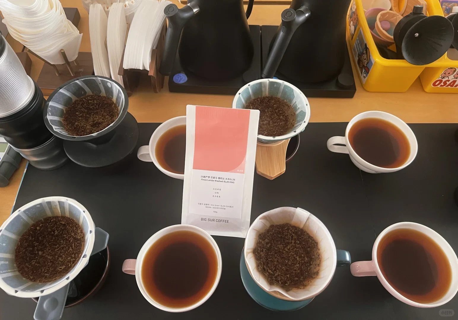 A bag of Big Sur Coffee stands among cups of brewed coffee and drippers filled with used grounds; kettles and paper filters are behind it.