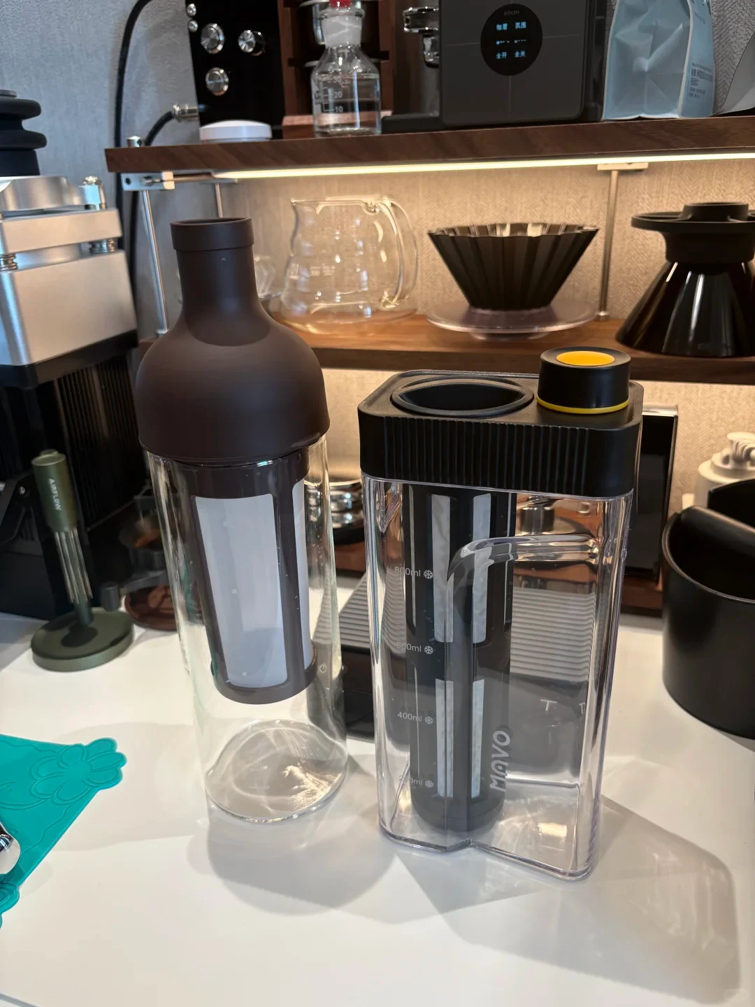 The owner’s HARIO wine-shaped coffee bottle stands beside a MAVO brick-shaped 800 ml brewer with a ribbed black lid and a yellow-ringed pour cap, on a counter in front of other coffee gear.