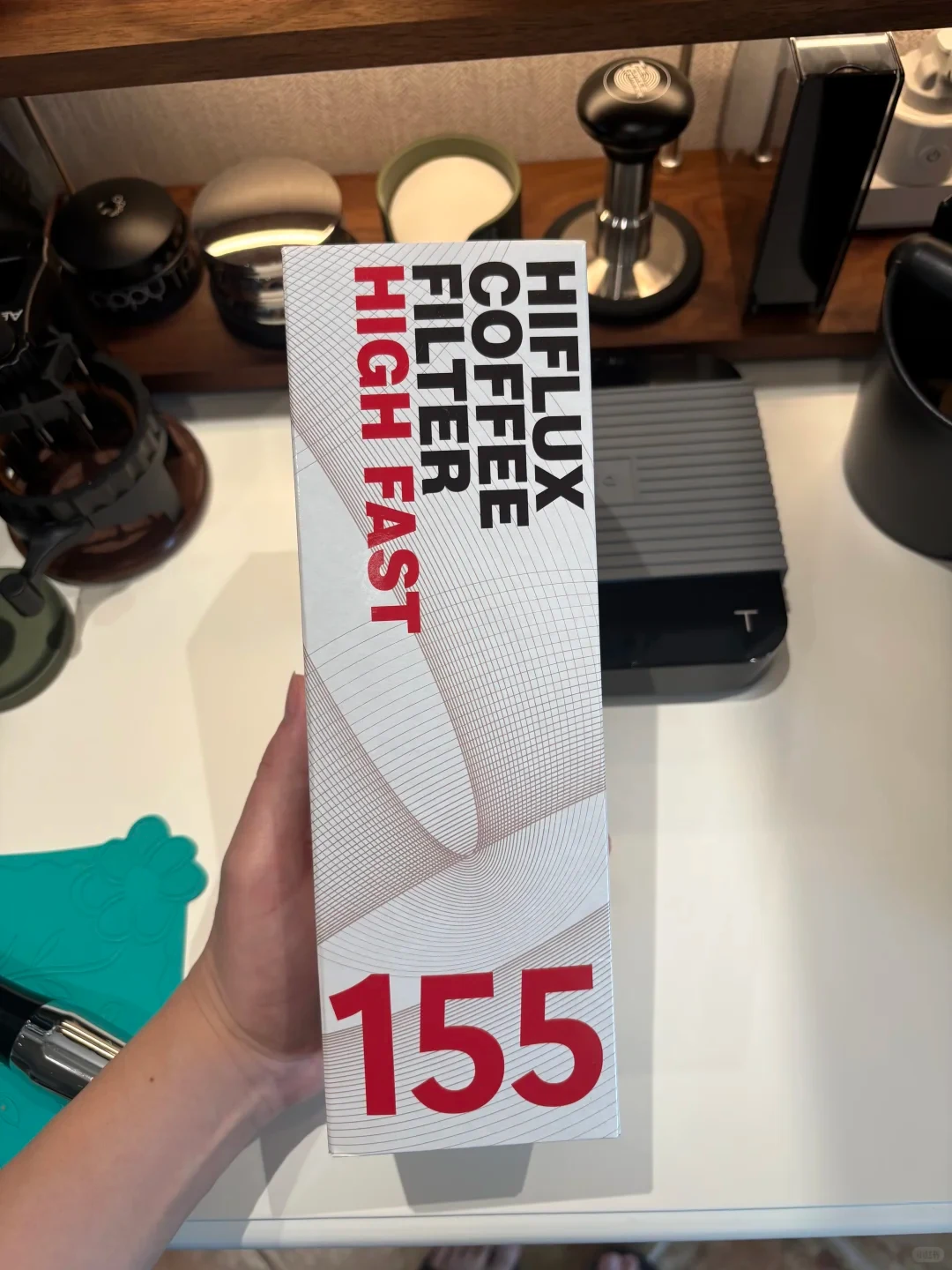 A hand holds a tall white box labeled “HIFLUX COFFEE FILTER,” “HIGH FAST,” and “155” above a countertop with coffee-making equipment.