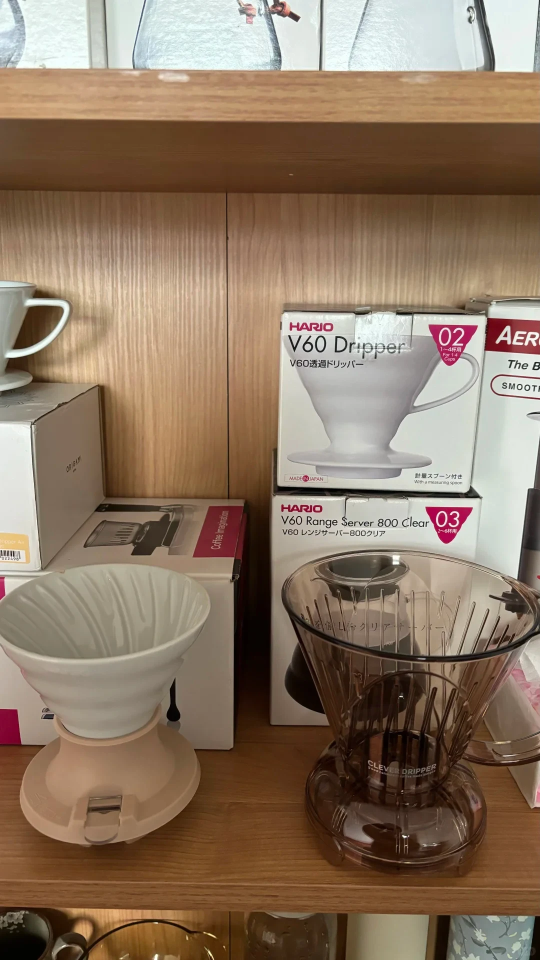 The owner’s shelf holds a white ceramic immersion dripper with a lever and a brown Clever Dripper. HARIO V60 and Range Server boxes and other coffee gear are in the background.