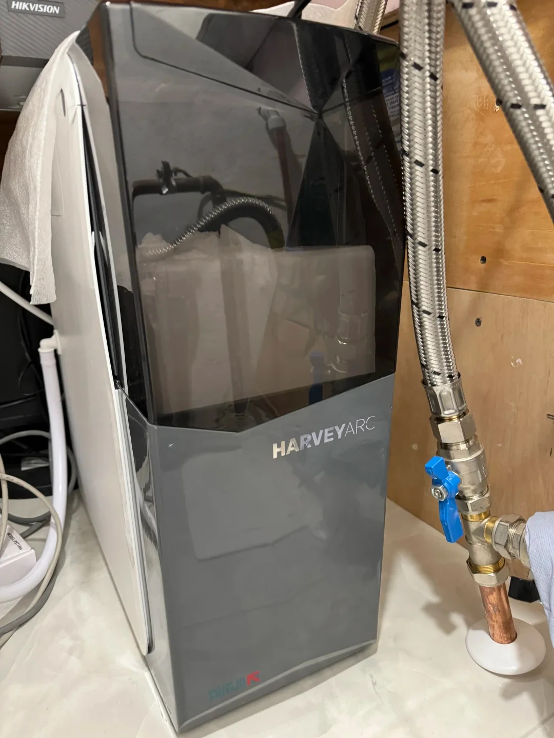 A Harvey Arc water softener installed beneath a counter beside stainless steel braided plumbing hoses and a blue shut-off valve.