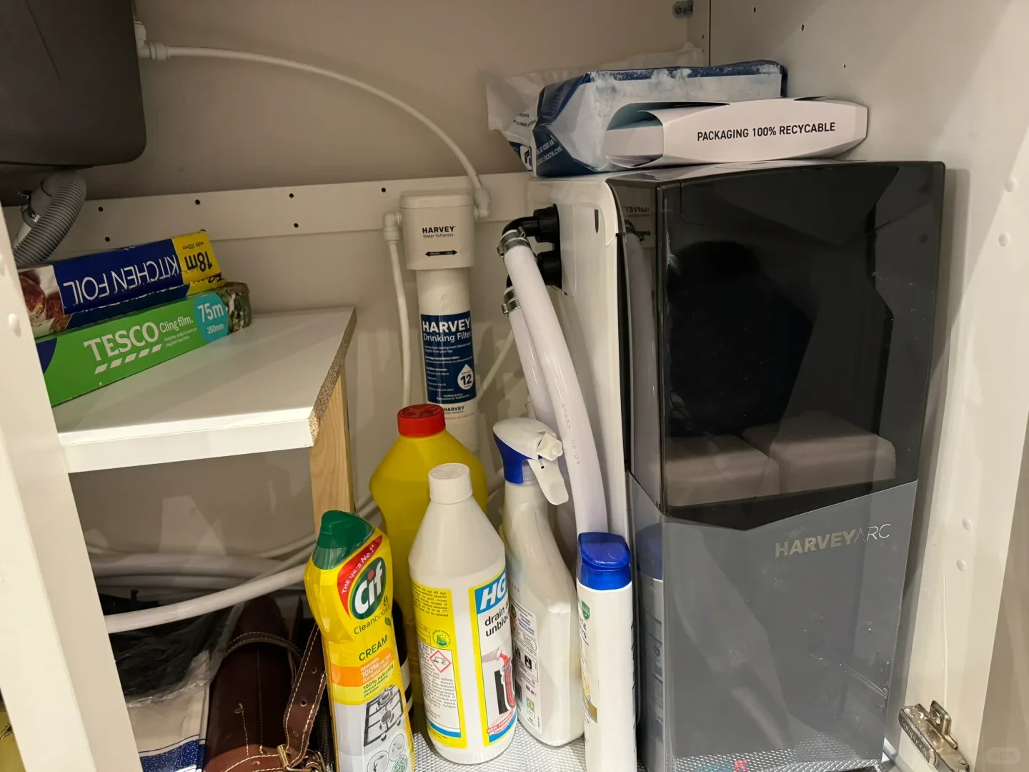 An open cabinet beneath a sink containing a Harvey Arc water softener system, a water filter, and various cleaning supplies such as Cif, HG drain unblocker, foil, and cling film.