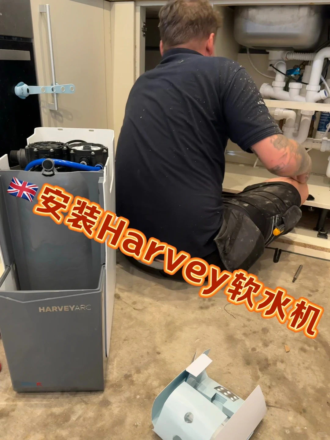 A technician sits on the floor working under a kitchen sink, next to an unboxed Harvey Arc water softener standing in the foreground.