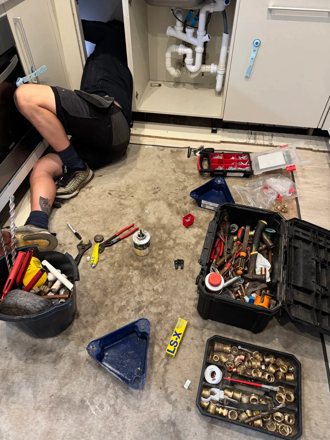 A plumber lies on a kitchen floor with his upper body inside an open lower cabinet fixing pipes beneath the sink, surrounded by toolboxes, plumbing fittings, wrenches, and sealant.