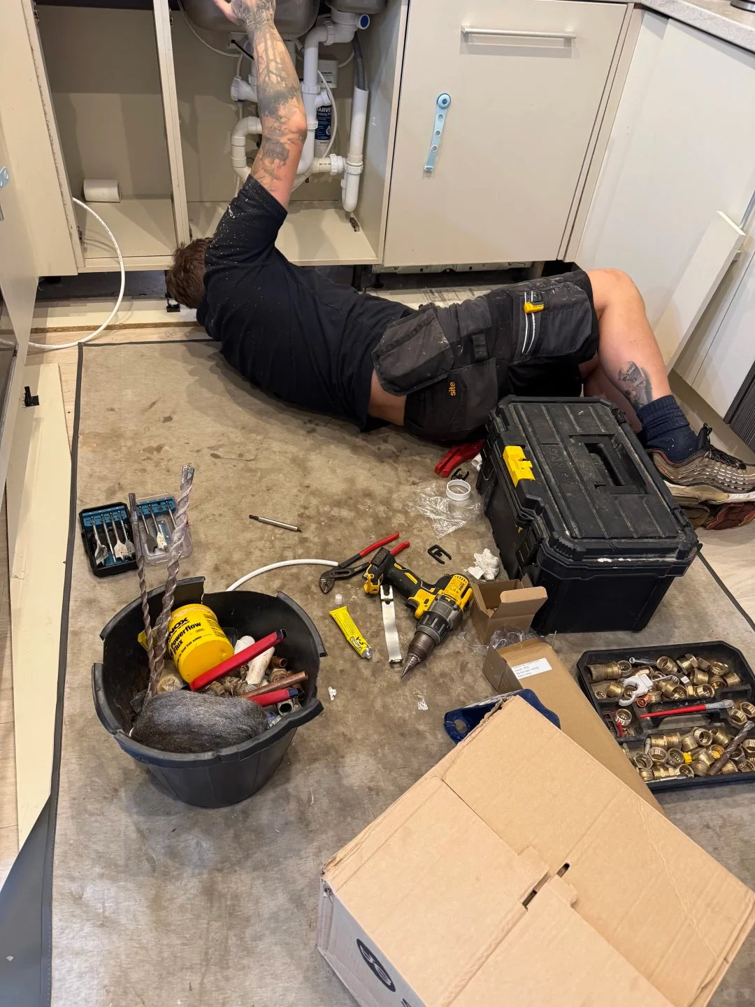A plumber lying on a protective mat on the floor working on plumbing pipes beneath an open kitchen cabinet, surrounded by various tools, a drill, a toolbox, and pipe fittings.