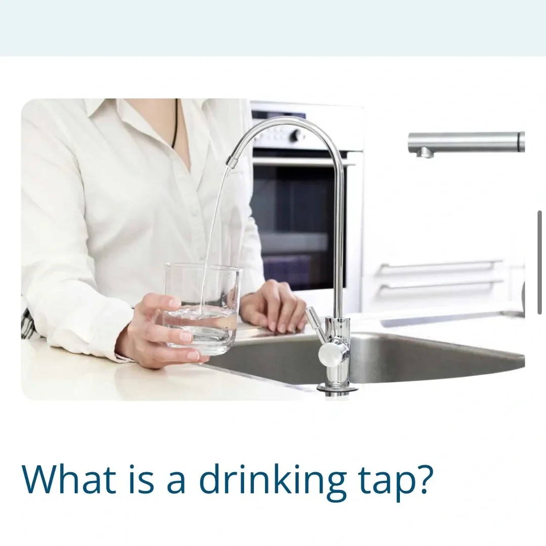 A person wearing a white collared shirt fills a clear drinking glass with water pouring from a slim, gooseneck chrome drinking water tap installed next to a kitchen sink. Modern kitchen cabinets and an oven appear blurred in the background. The bold text below the photo asks, 'What is a drinking tap?'