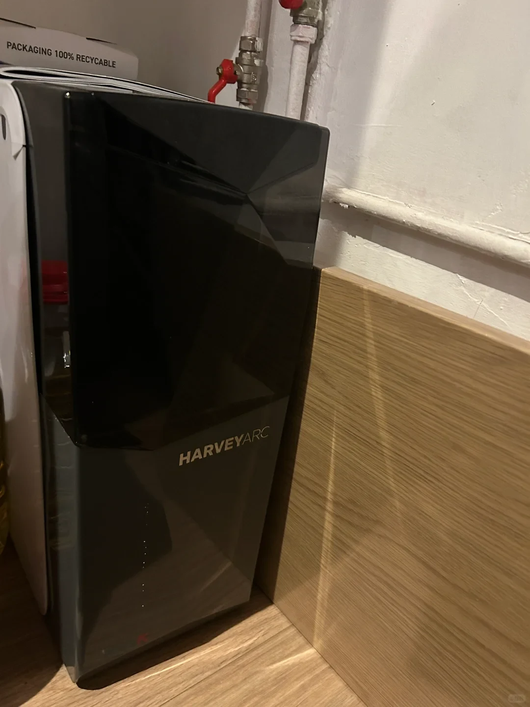 A black Harvey Arc water softener standing upright on a light wooden floor next to a wooden partition, with plumbing pipes and a red valve visible on the wall behind it.