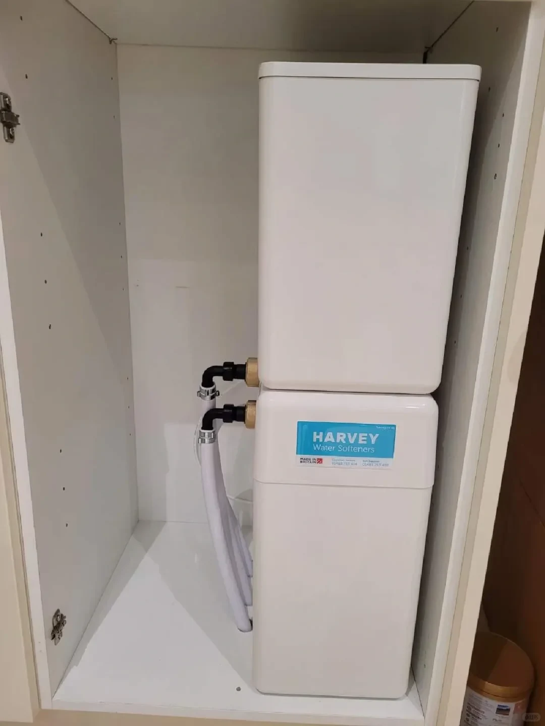 A white Harvey Water Softeners appliance installed vertically inside a white cabinet, connected with plumbing pipes and hoses on its left side.