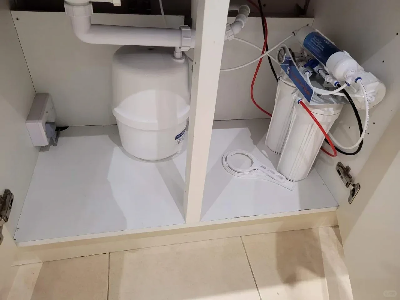 An under-sink reverse osmosis water filtration system installed inside a white kitchen cabinet, showing a white pressure tank on the left and vertical filter canisters, tubing, and a filter wrench on the right.