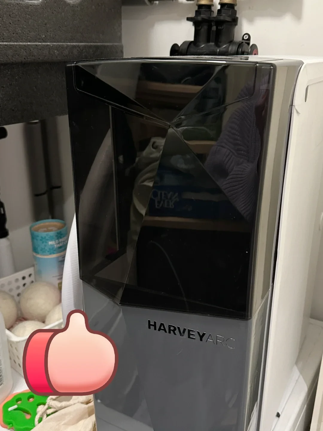 A Harvey Arc water softener installed in a utility space, featuring a dark geometric faceted front panel over a grey base labeled HARVEYARC, with plumbing valves on top and a thumbs-up sticker covering the lower left.