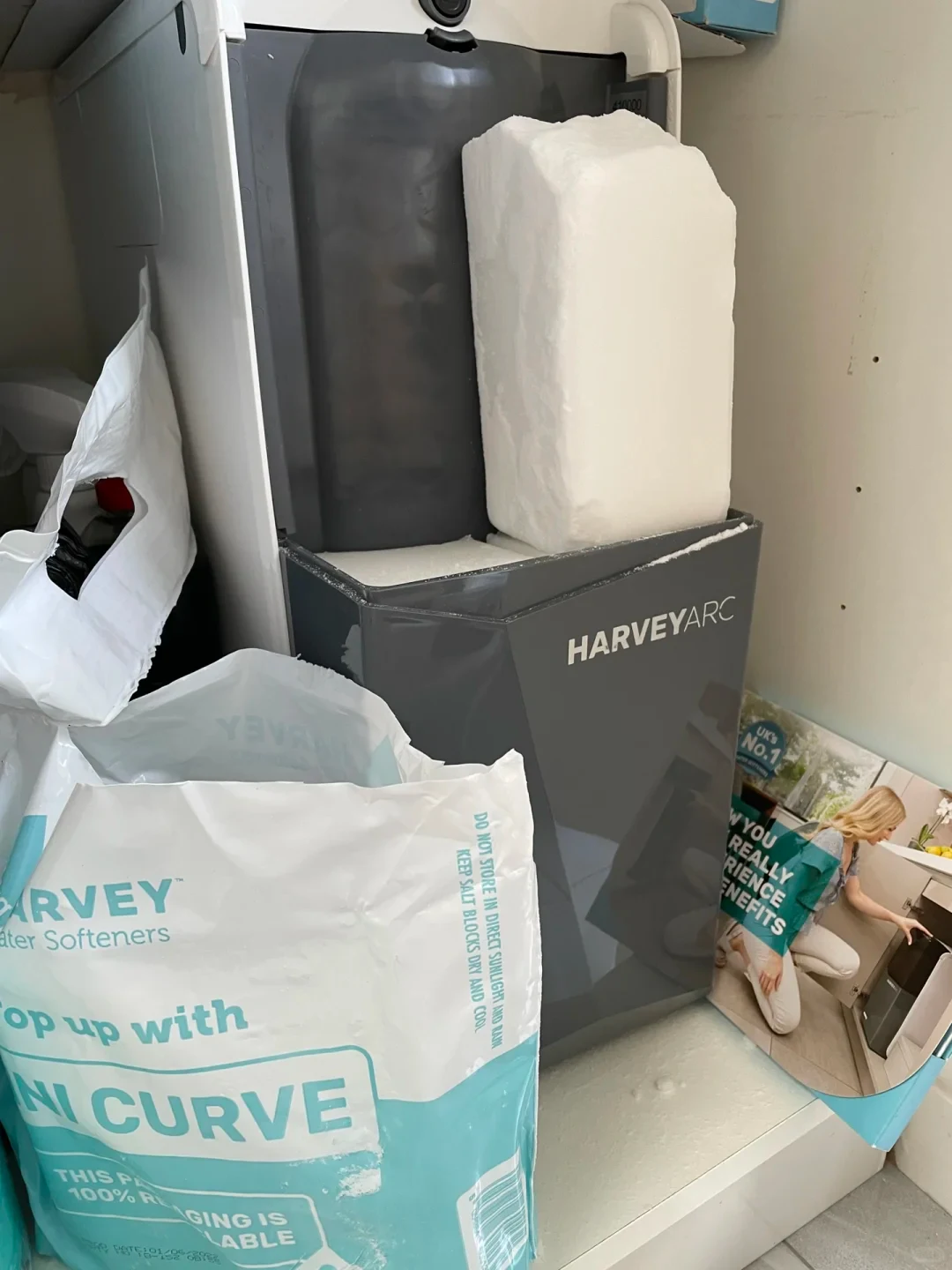 A HarveyArc water softener installed inside a cabinet, with an upright block of white salt resting inside the open compartment. Next to it stands a branded promotional brochure, and an open bag of Harvey Mini Curve salt blocks rests in the foreground.