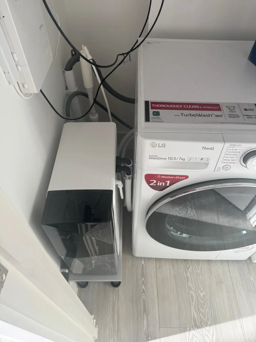A top-down view of a laundry area with a white LG 2-in-1 front-loading washer-dryer unit next to a compact water softener or filtration appliance on a wheeled tray, with associated plumbing and wires along the wall.