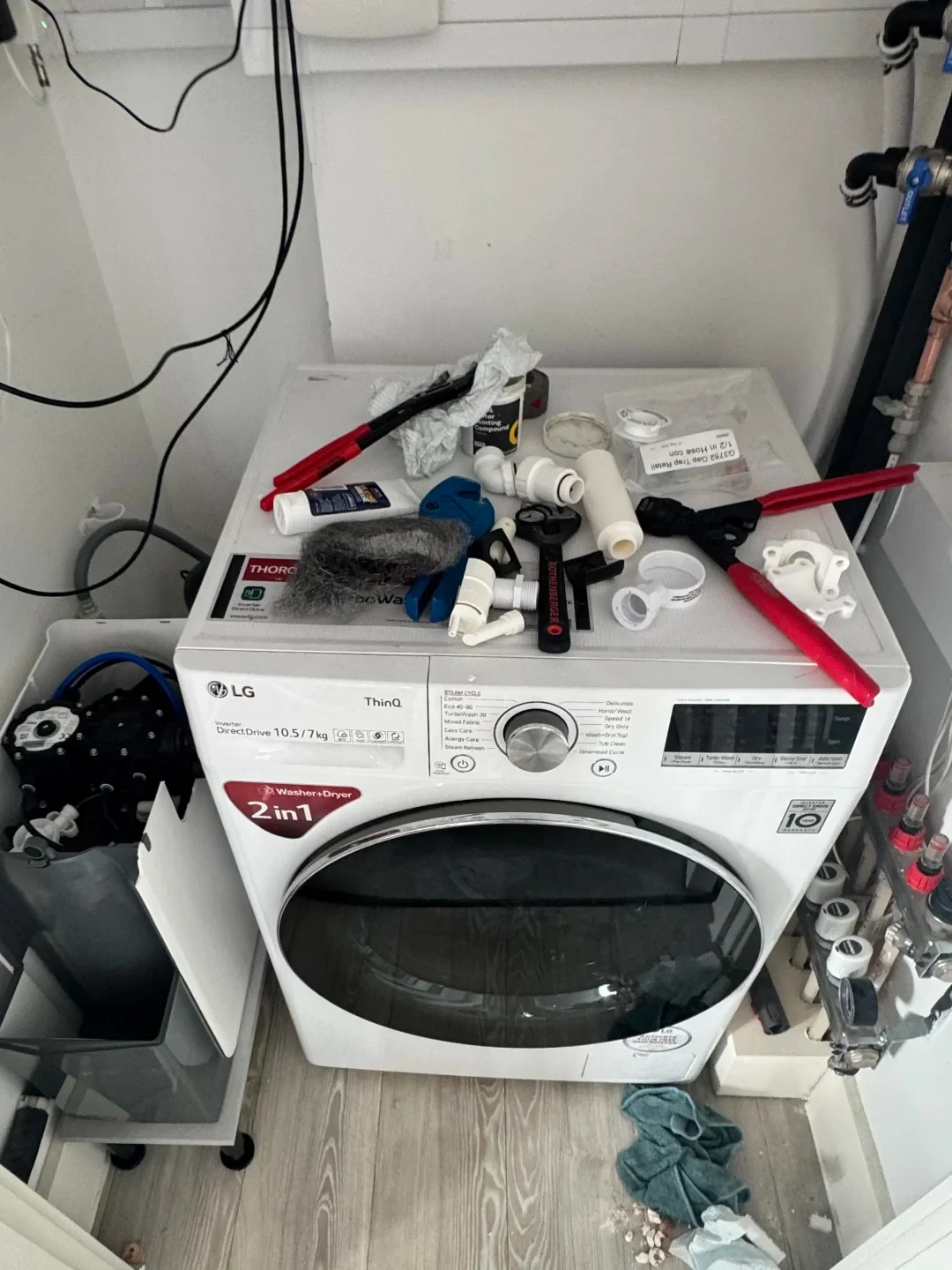 A front-loading white LG ThinQ 2-in-1 washer and dryer set in a small utility closet with plumbing fixtures to the right and an open bin to the left. The top of the washing machine is covered in various plumbing tools, plastic pipe fittings, tubes, wire wool, and red-handled pipe cutters, indicating ongoing installation or repair work.