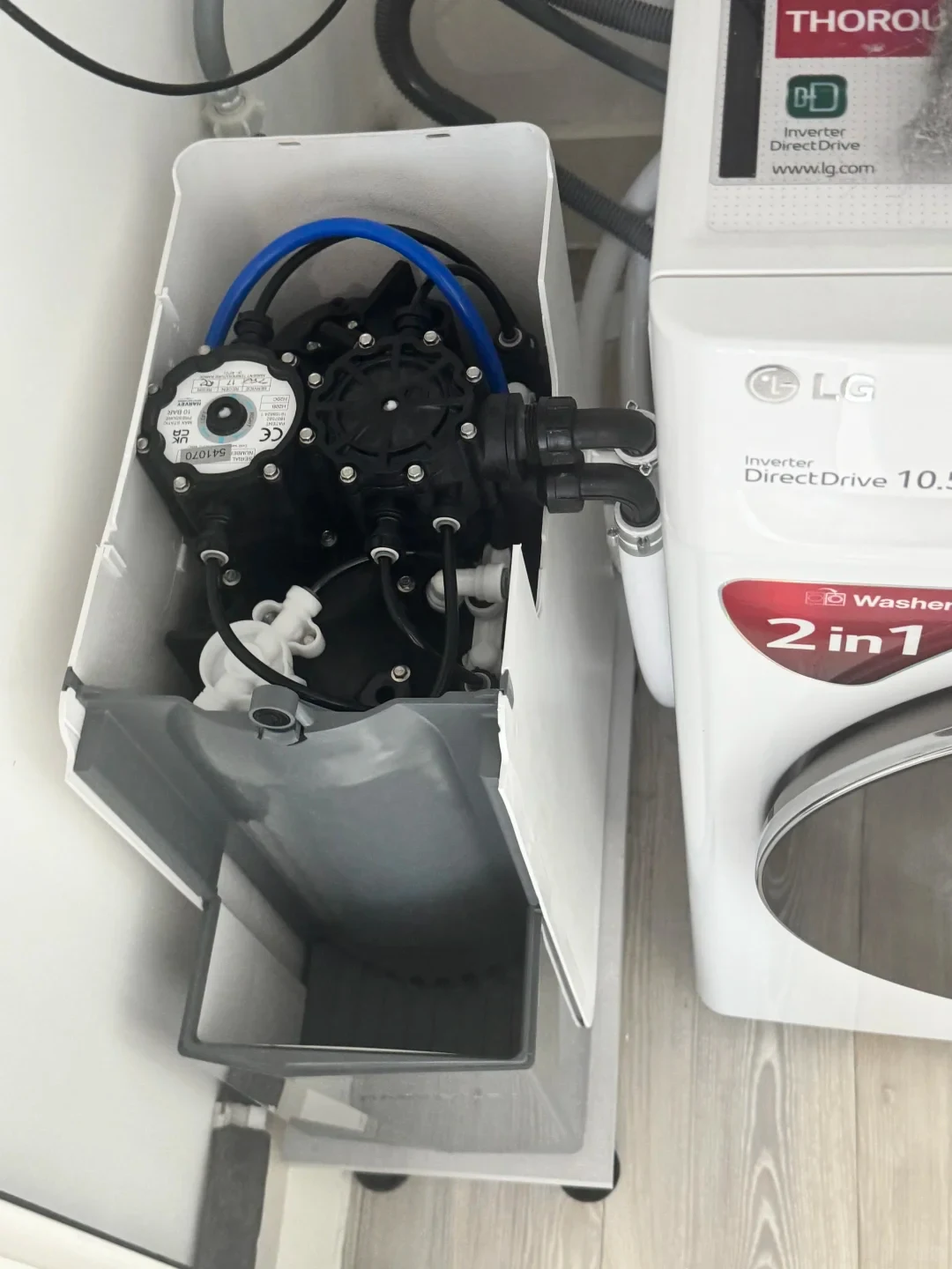 A top-down view of an opened compact water softener unit next to a white LG washing machine, showing the internal valves, water meter, tubes, and salt compartment.