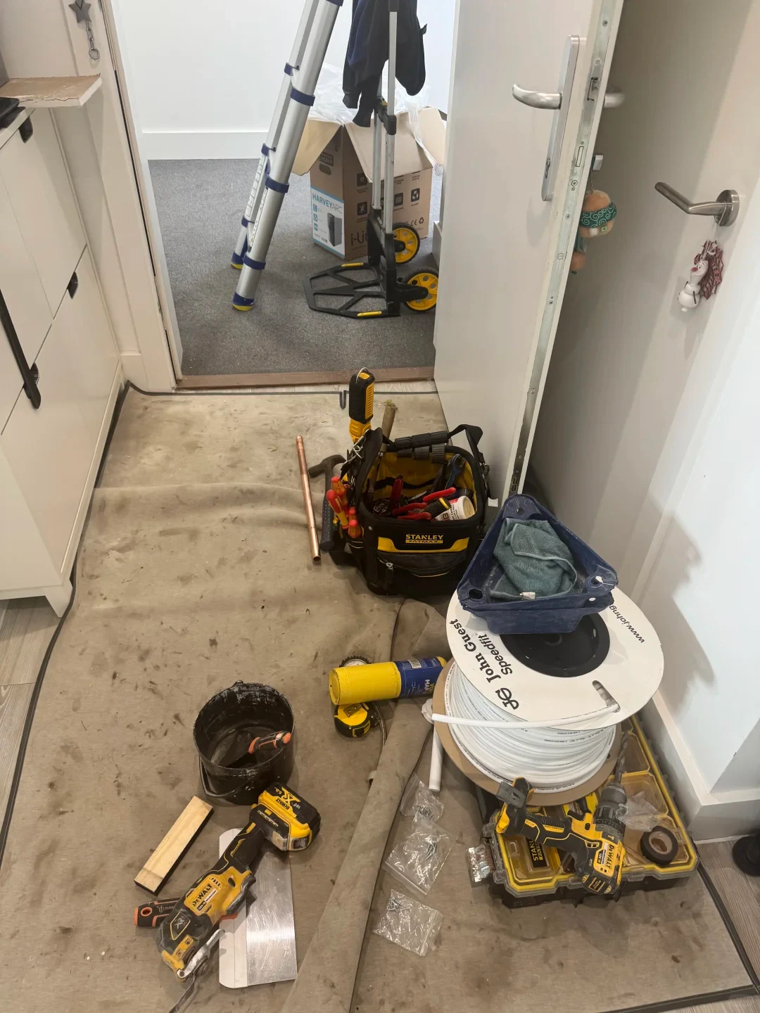 A floor covered with a protective drop cloth during renovation or plumbing work. Scattered across the floor are various tools and supplies, including a yellow and black Stanley tool bag filled with hand tools, DeWalt power tools, a roll of white John Guest Speedfit tubing, a small black bucket, a copper pipe, and screws in plastic bags. An open doorway in the background reveals a carpeted room with an extended telescoping ladder, a hand truck, and a cardboard box.