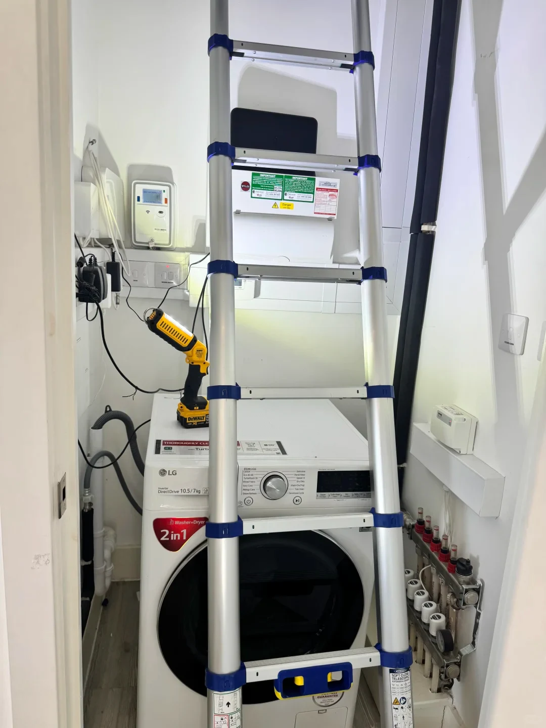 A narrow utility closet containing a white LG front-loading washer-dryer combo, with a silver telescopic aluminum ladder set up in front of it extending upward. A yellow DeWalt cordless work light sits on top of the washing machine, illuminating the space, while an underfloor heating manifold lines the lower right wall.