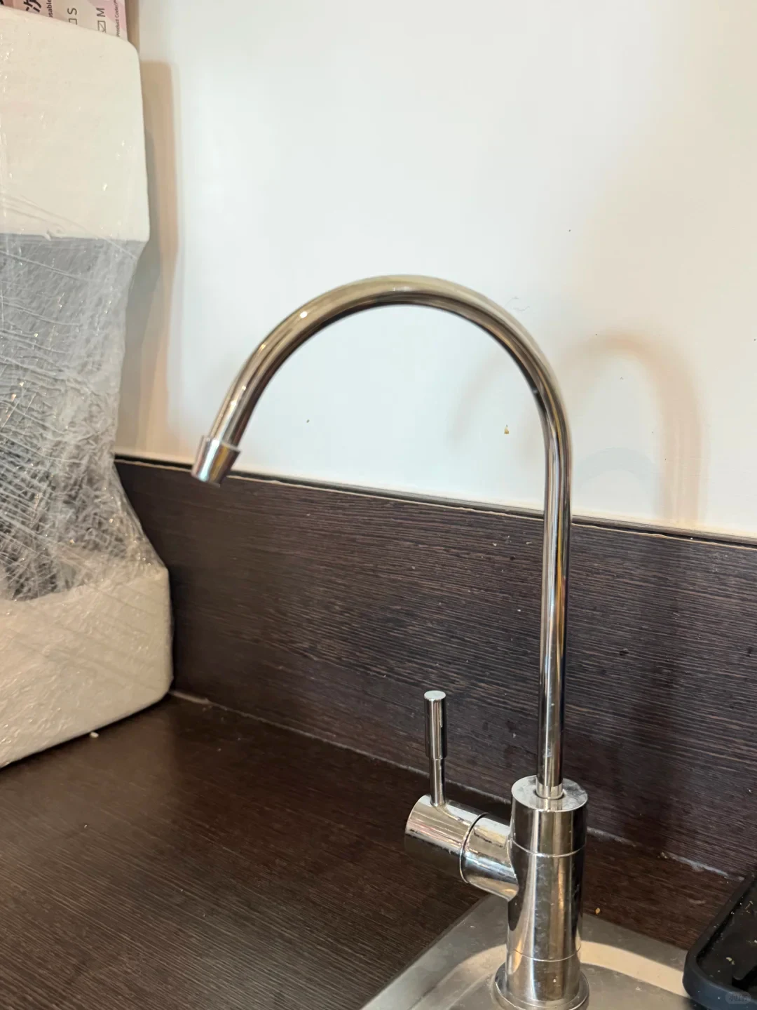 A polished chrome gooseneck drinking water faucet mounted on the edge of a stainless steel sink beside a dark wood backsplash and white wall.
