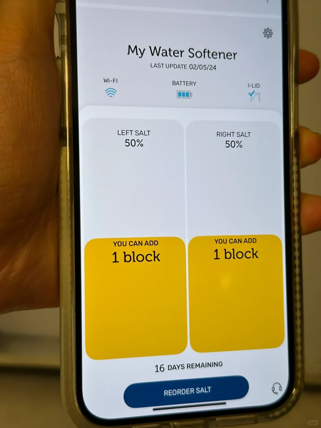 A hand holding a smartphone showing a smart water softener app interface with left and right salt levels at 50 percent, indicating one block can be added for each.