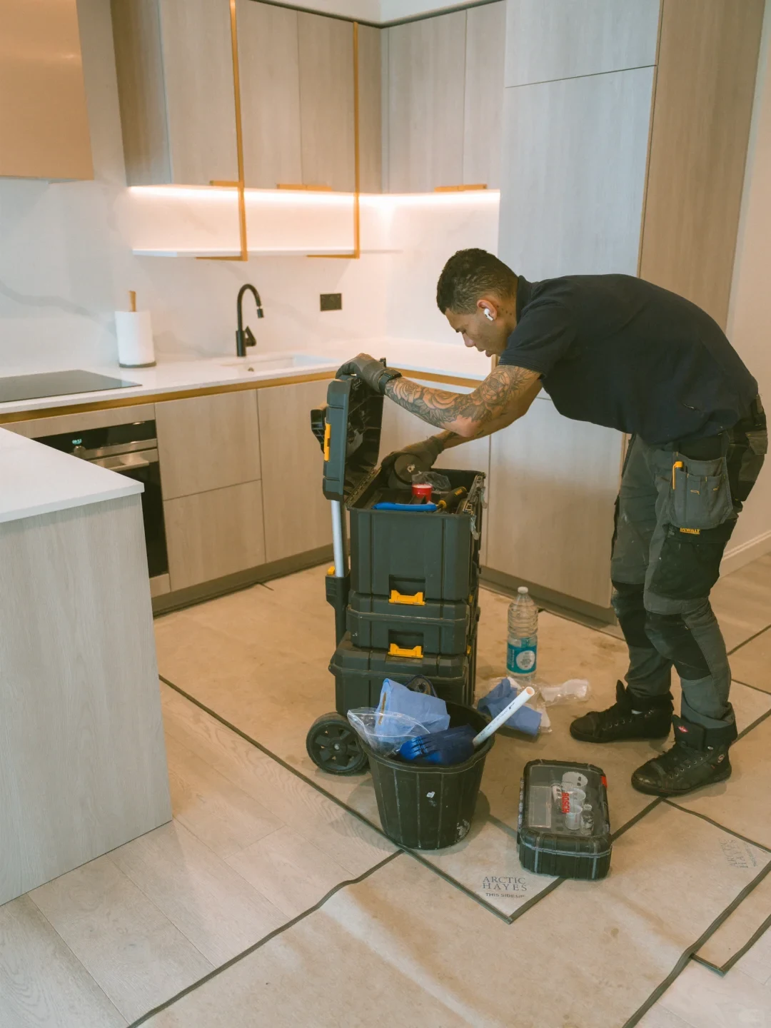 A male tradesperson with tattooed arms stands inside a modern kitchen with light wood cabinetry, leaning over to open a stacked, wheeled black and yellow modular toolbox. Additional tools, a bucket, and a tool case rest on a protected section of the tile floor beside him.