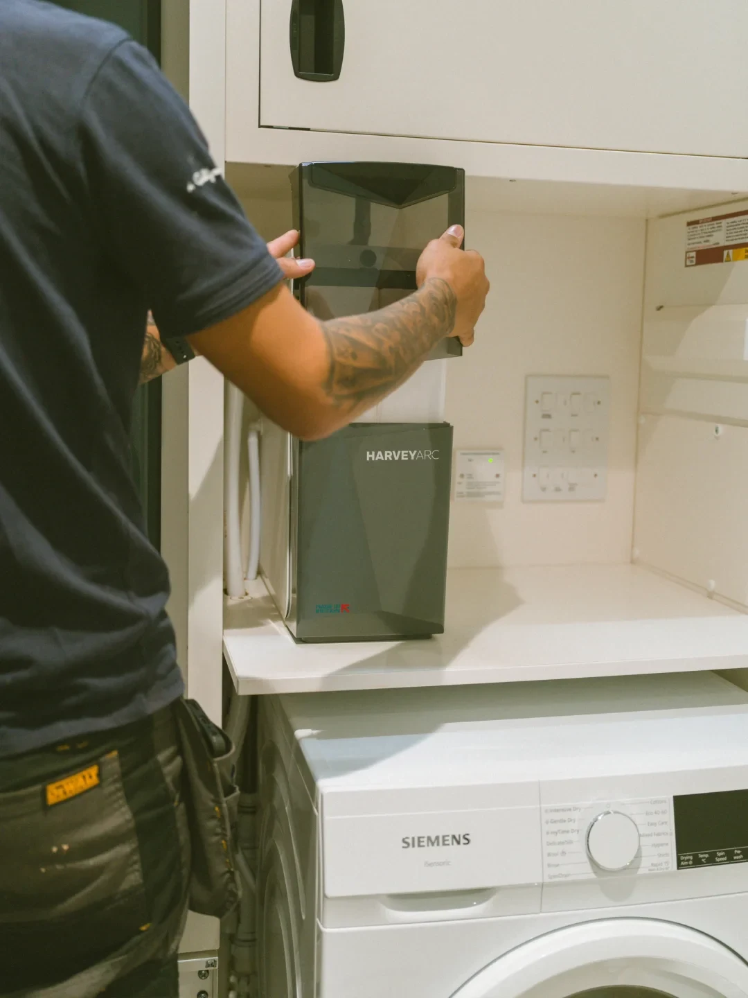 A technician with tattooed arms handles a grey Harvey Arc water softener installed on a shelf inside a utility cupboard, situated above a white Siemens front-load washing machine.