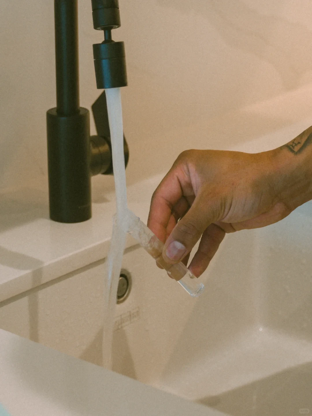 A person's hand holding a small transparent glass vial under a stream of running water from a modern matte black faucet over a white sink.