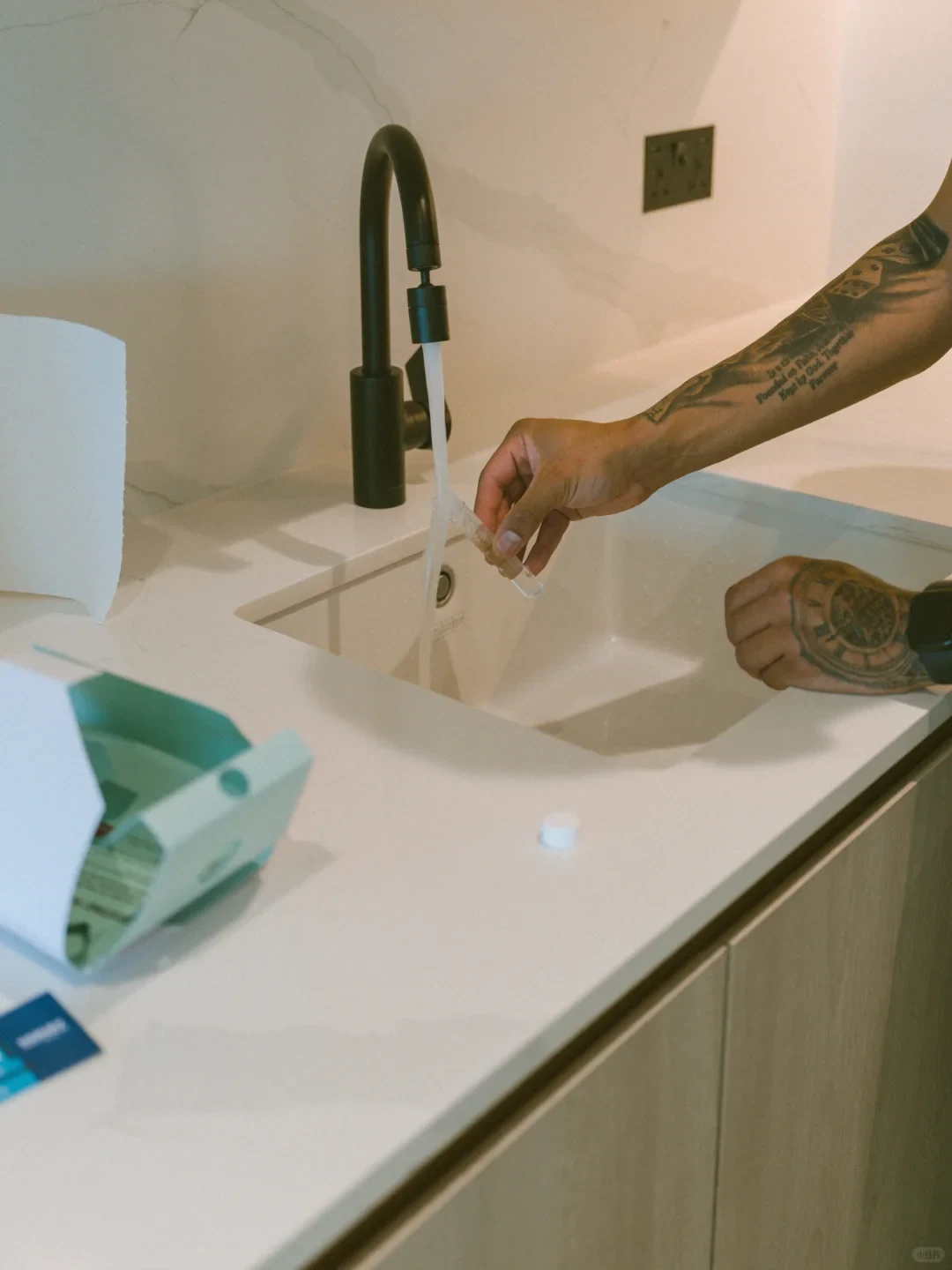 A person with tattooed arms holds a small clear vial under running water from a sleek black gooseneck faucet over a white countertop sink, with an open light teal product box and a white cap resting on the counter.