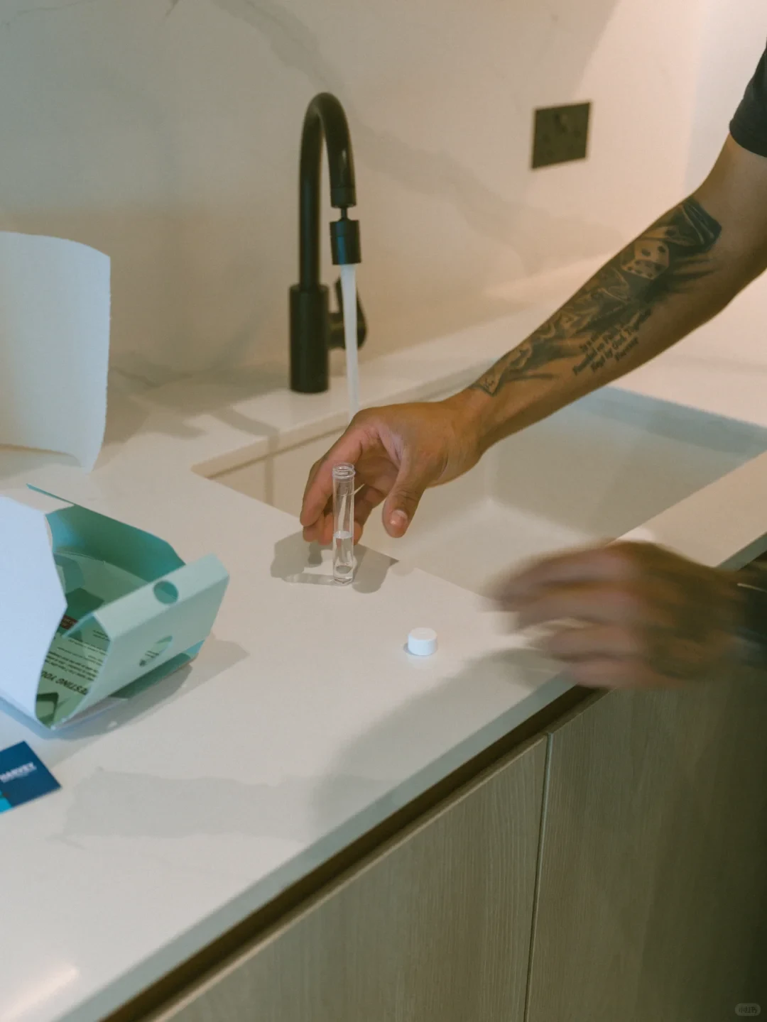 A person with tattooed arms standing by a kitchen sink with running water from a black faucet, handling a small clear test vial on the white countertop beside an opened water testing kit box.