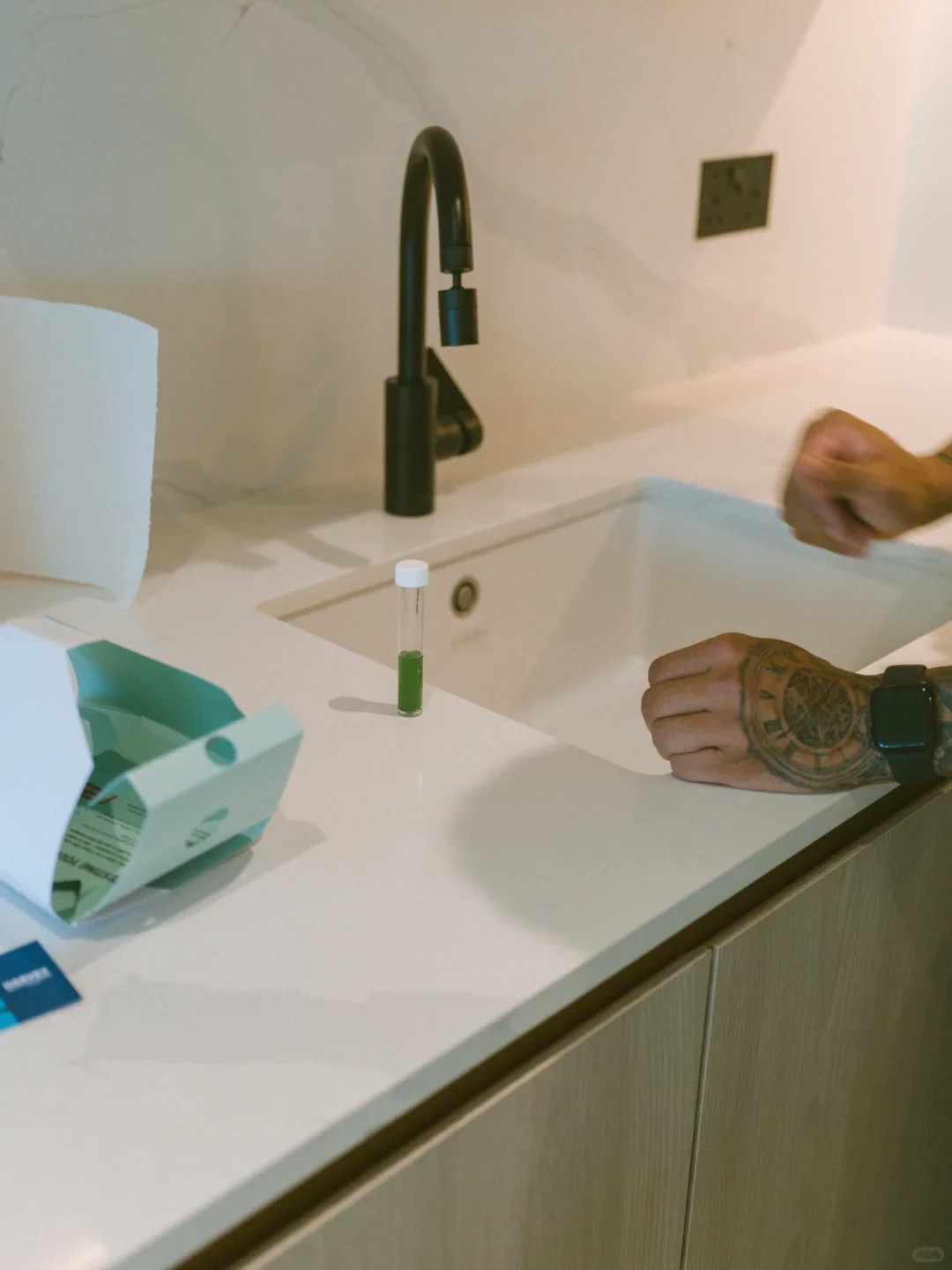 A tattooed person stands beside a white kitchen sink where a small test tube containing green liquid stands upright on the white countertop next to an open testing kit box and a matte black faucet.