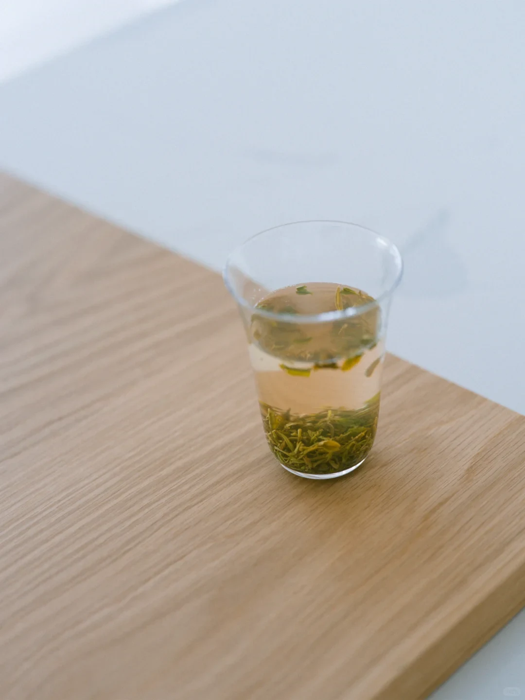 A clear glass cup filled with brewed green tea, showing tea leaves both floating on the surface and resting at the bottom, placed on a light-colored wooden table surface.