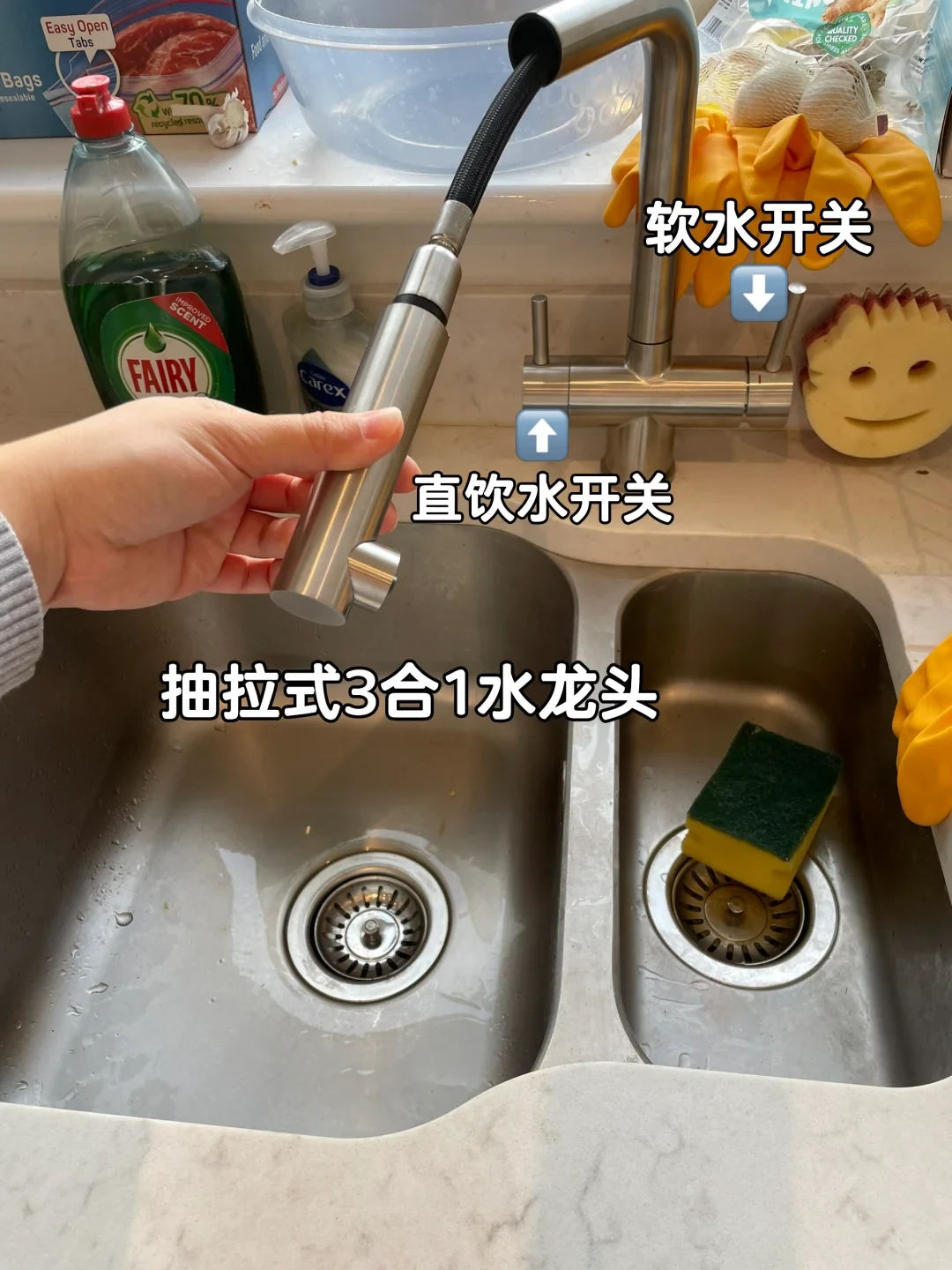 A person holding the pull-out sprayer head of a stainless steel 3-in-1 kitchen faucet positioned over a double-basin sink.