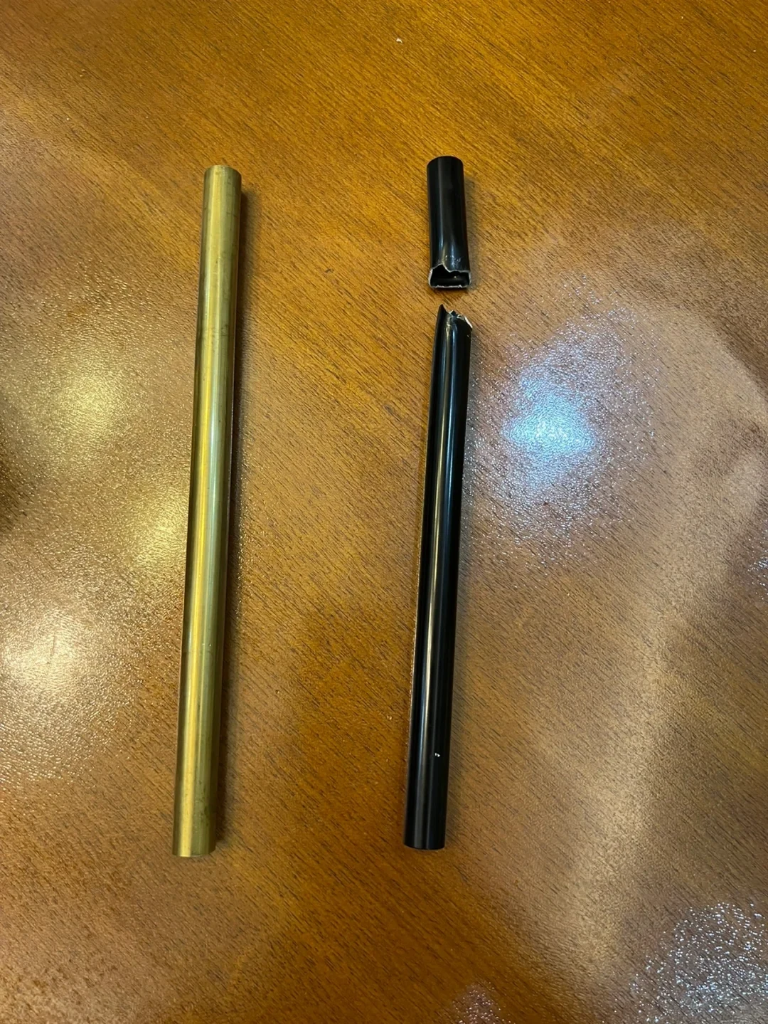 A gold-colored brass rod lies next to a black tube broken into two pieces on a wooden tabletop.