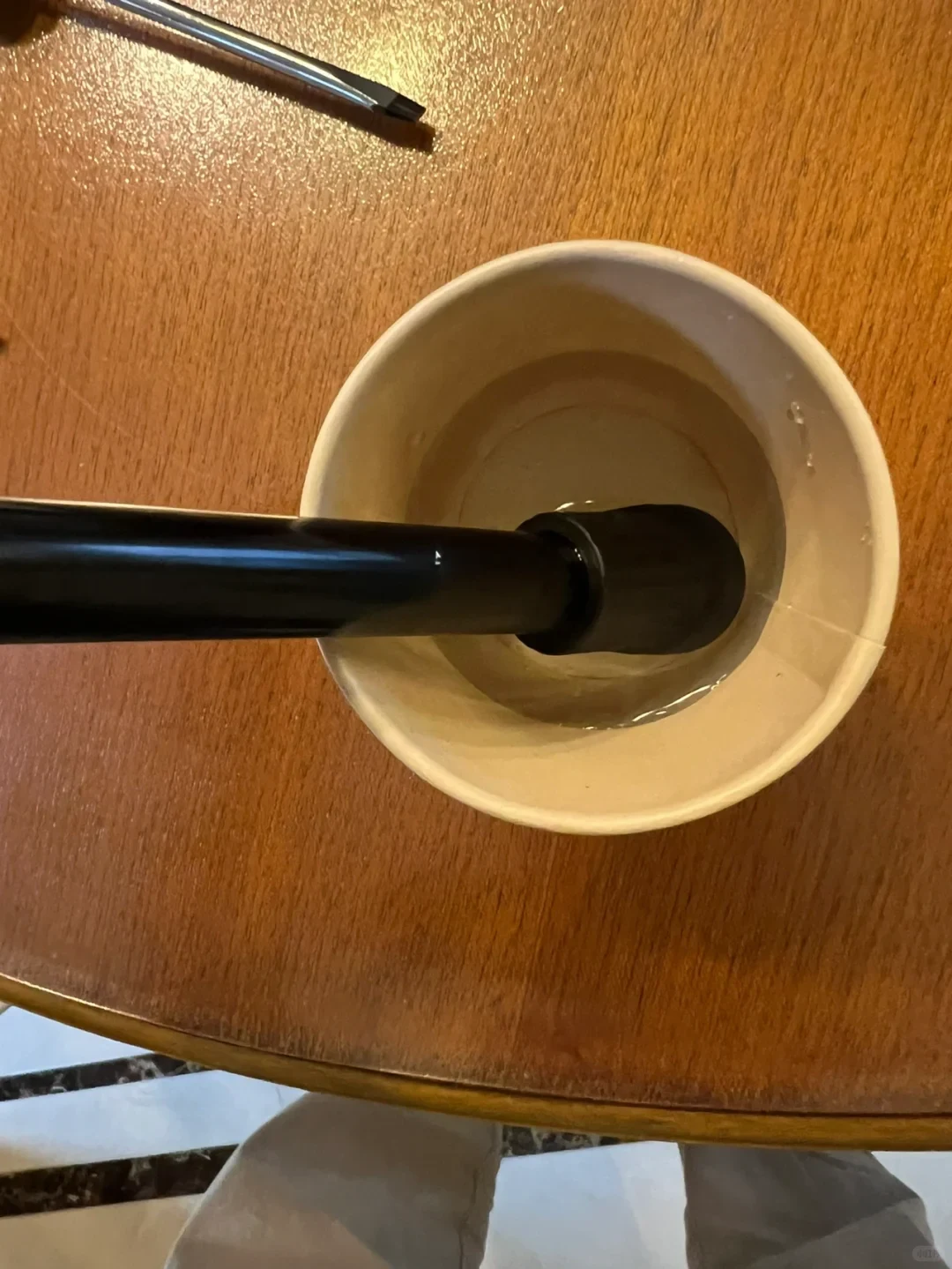 Top-down view of a black tube with an attached nozzle submerged inside a paper cup containing clear liquid on a wooden tabletop, with a flathead screwdriver visible at the top.
