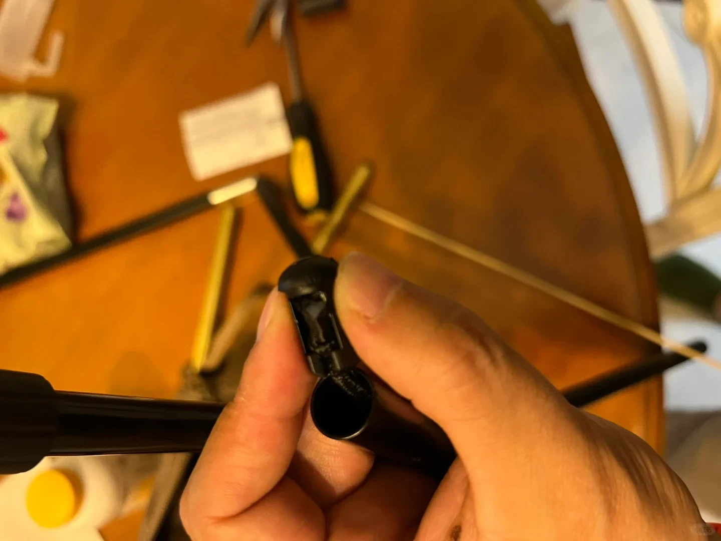 A close-up of a person holding a damaged black plastic connector pulled out from the opening of a hollow black metal pole, with tools and disassembled rods on a wooden table in the background.