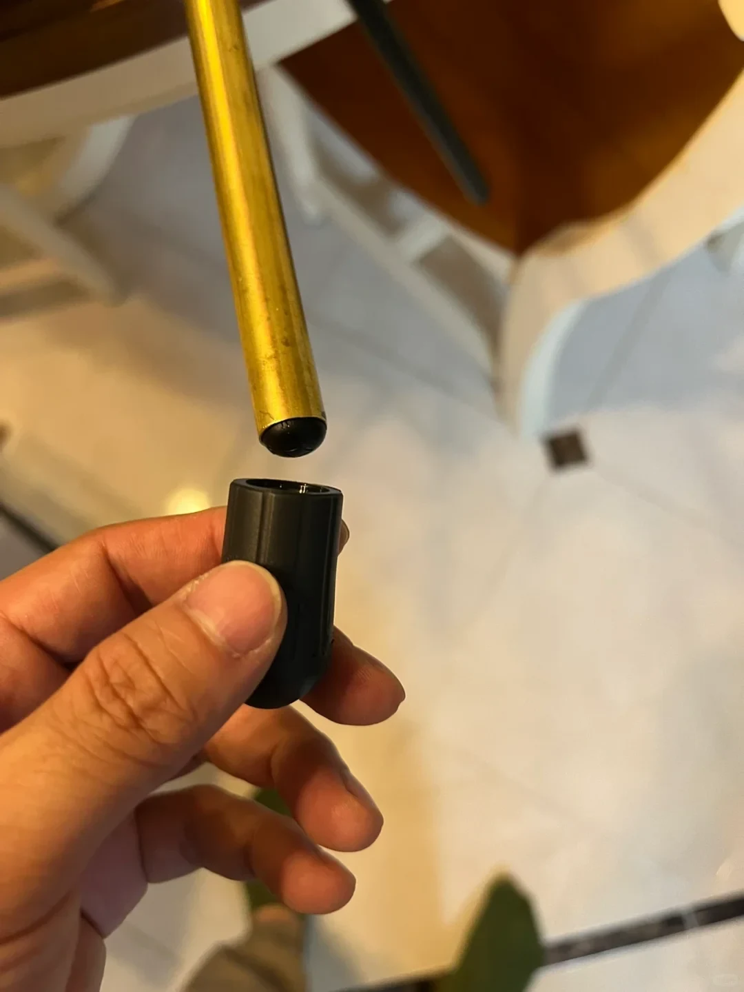 A person's hand holding a black rubber foot cap positioned just below the black-tipped end of a gold-colored metal chair or furniture leg.