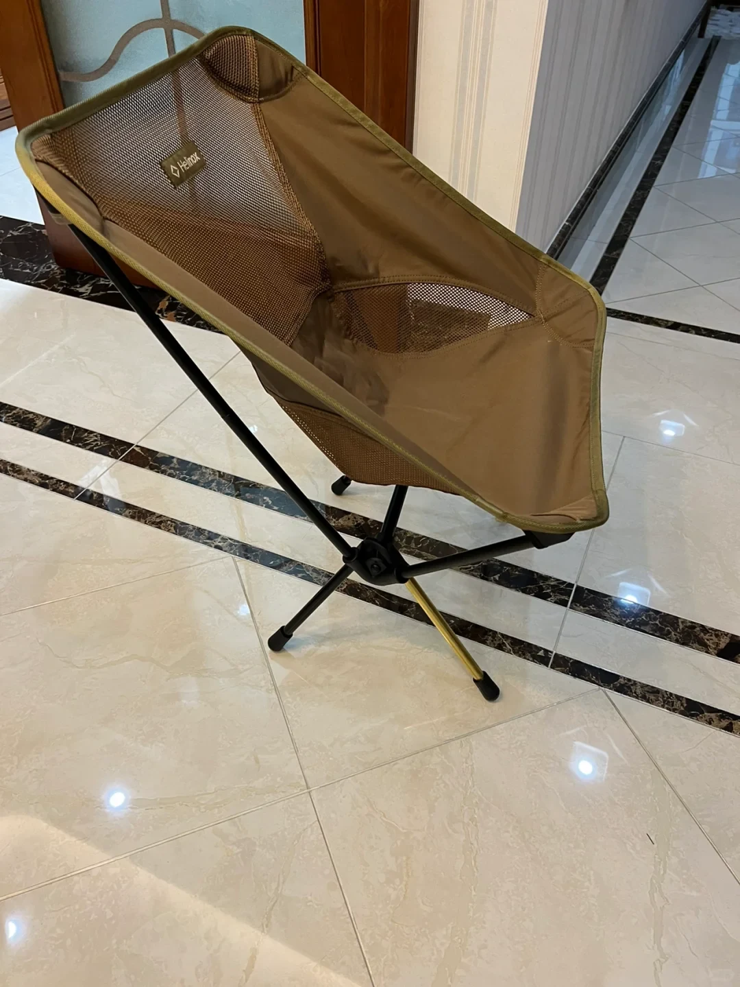 An assembled tan Helinox folding camp chair with mesh panels placed on a tiled indoor floor.