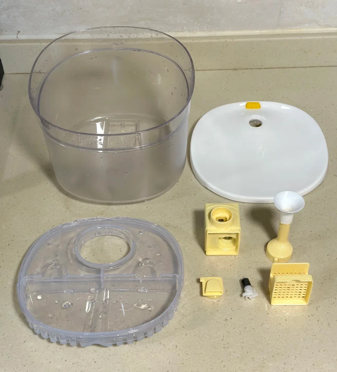 Disassembled parts of a pet water fountain arranged on a beige countertop, including a clear plastic reservoir, a white top lid, an oval clear tray, a pump housing, a water pipe spout, an impeller, and small plastic filter accessories.